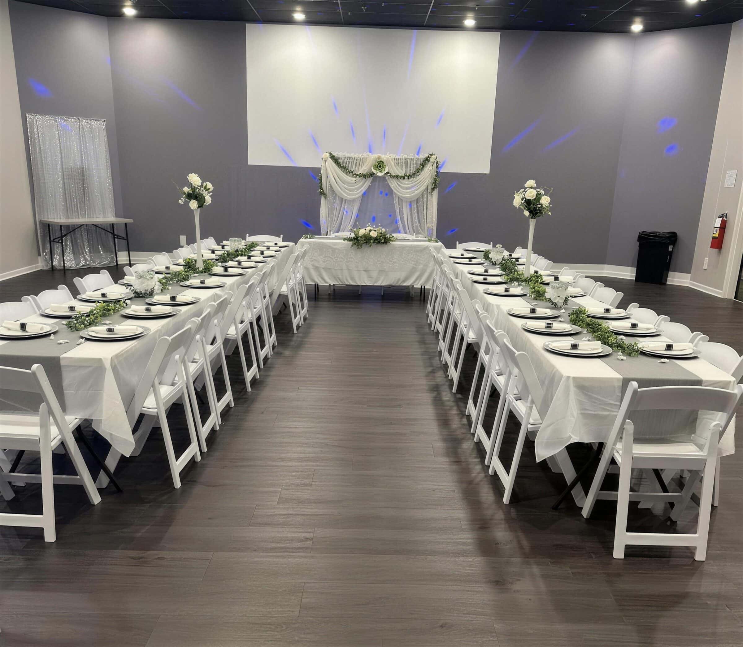 U-shaped tables with sage runners, black chargers and tall white rose vases facing a draped sweetheart table with blue uplighting at The Cove at St. Johns, Fruit Cove FL