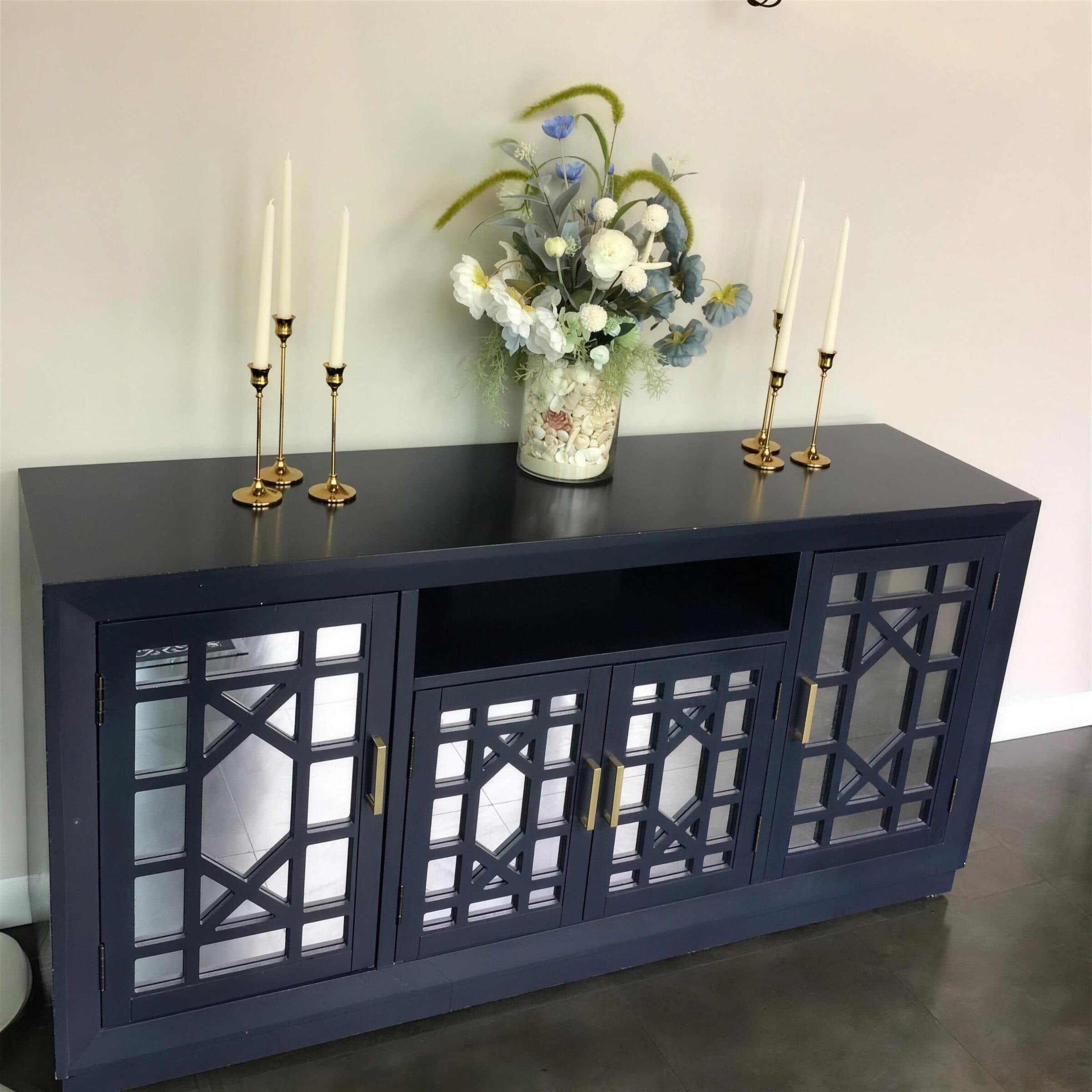 Navy mirrored sideboard styled with five gold taper candlesticks and a white, blue and green silk arrangement in a shell-filled glass vase at The Cove at St. Johns, Fruit Cove FL