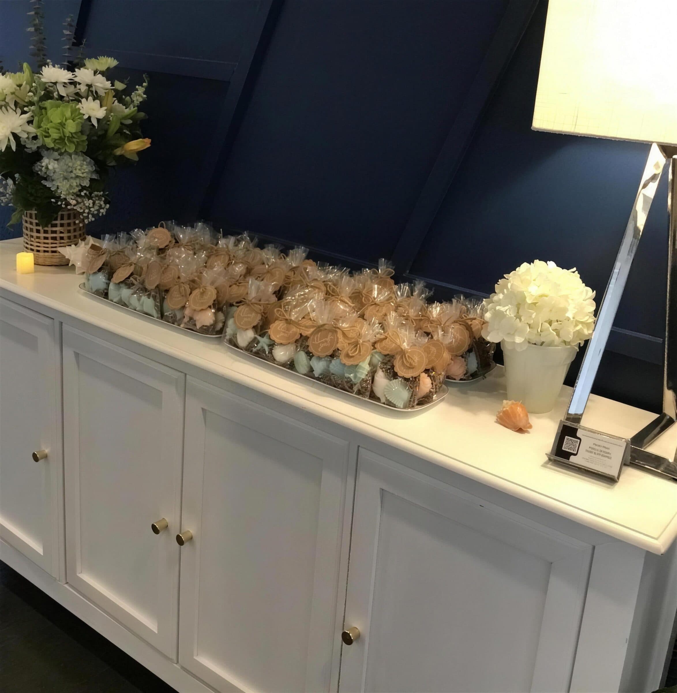 Trays of seashell soap favors in cellophane with kraft thank-you tags on a white credenza, white and green florals beside them, at The Cove at St. Johns, St. Johns County FL