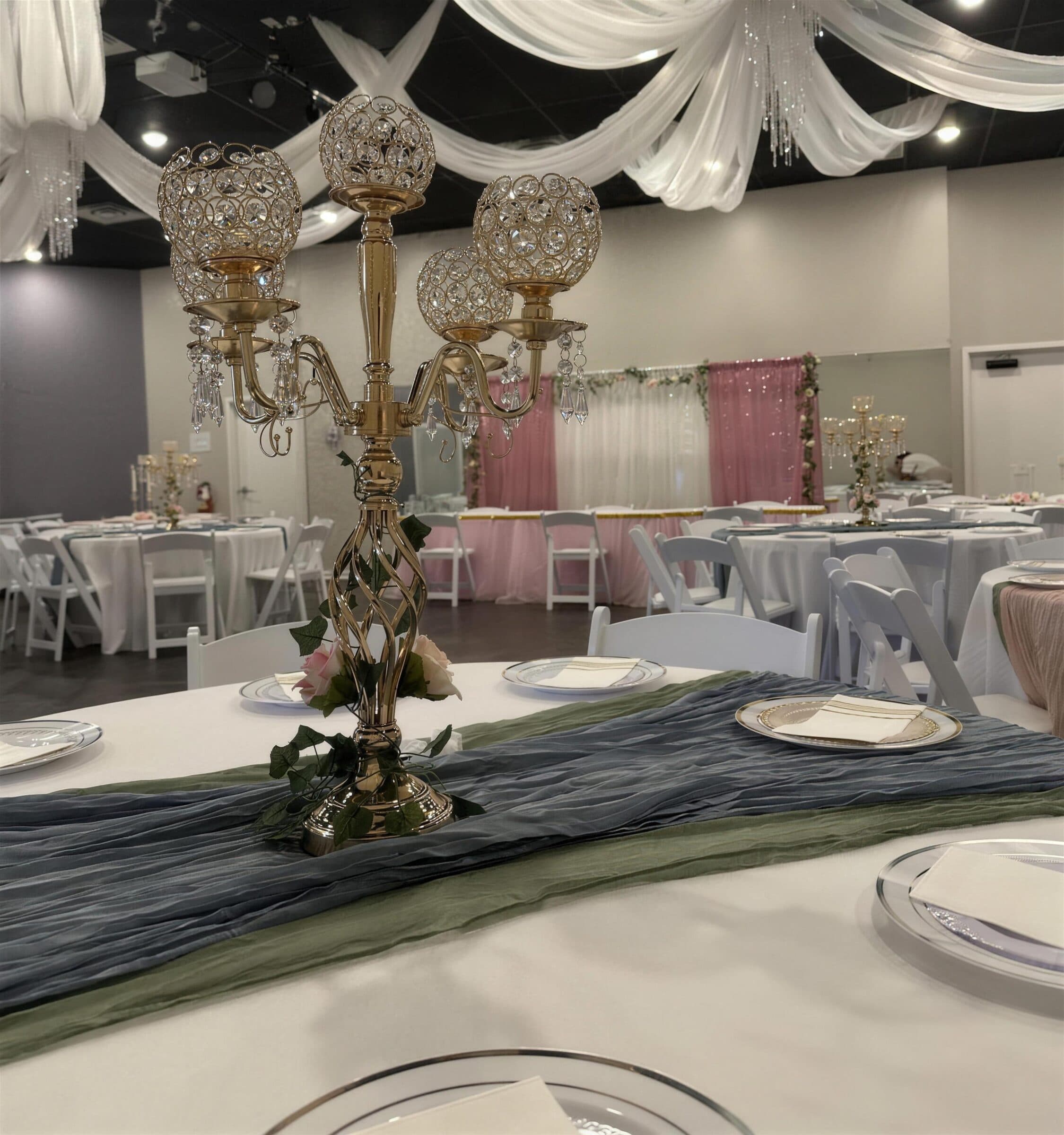 Gold crystal-globe candelabra centerpiece with pink roses and ivy on dusty blue and sage runners, white ceiling draping overhead, at The Cove at St. Johns, Fruit Cove FL