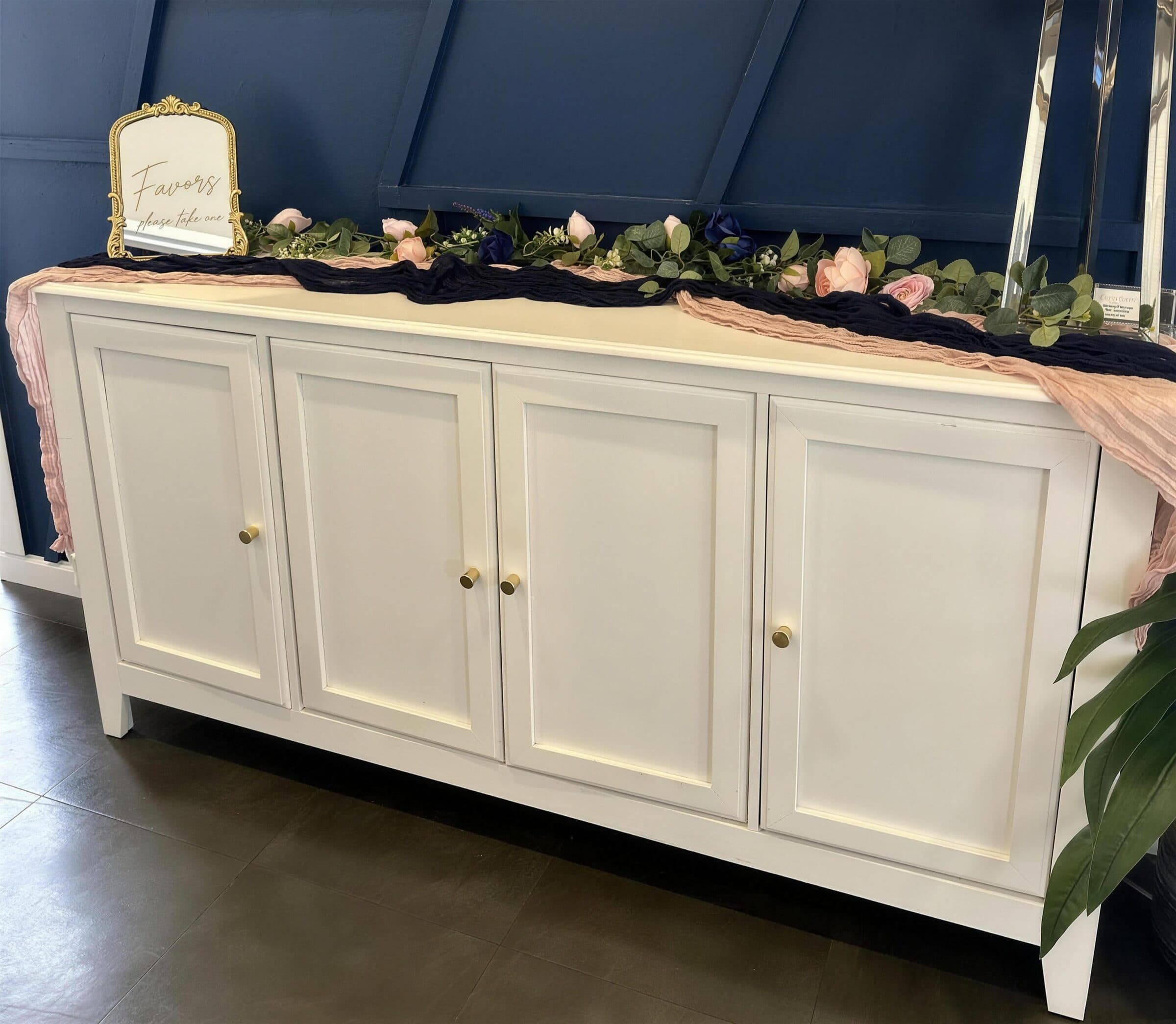 Gold-framed Favors sign on a white sideboard dressed with navy and blush cheesecloth and a garland of pink roses, navy blooms and eucalyptus at The Cove at St. Johns, near Jacksonville, FL