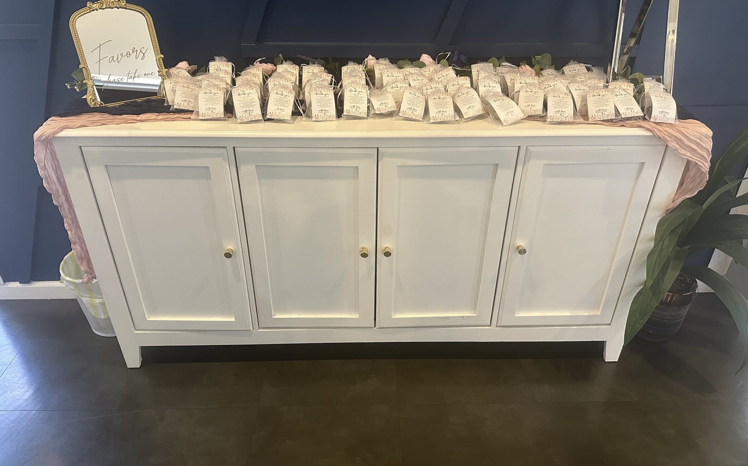 Rows of clear favor bags with floral thank-you tags lined across the same white sideboard under a blush runner and gold Favors sign at The Cove at St. Johns, Fruit Cove, St. Johns County FL