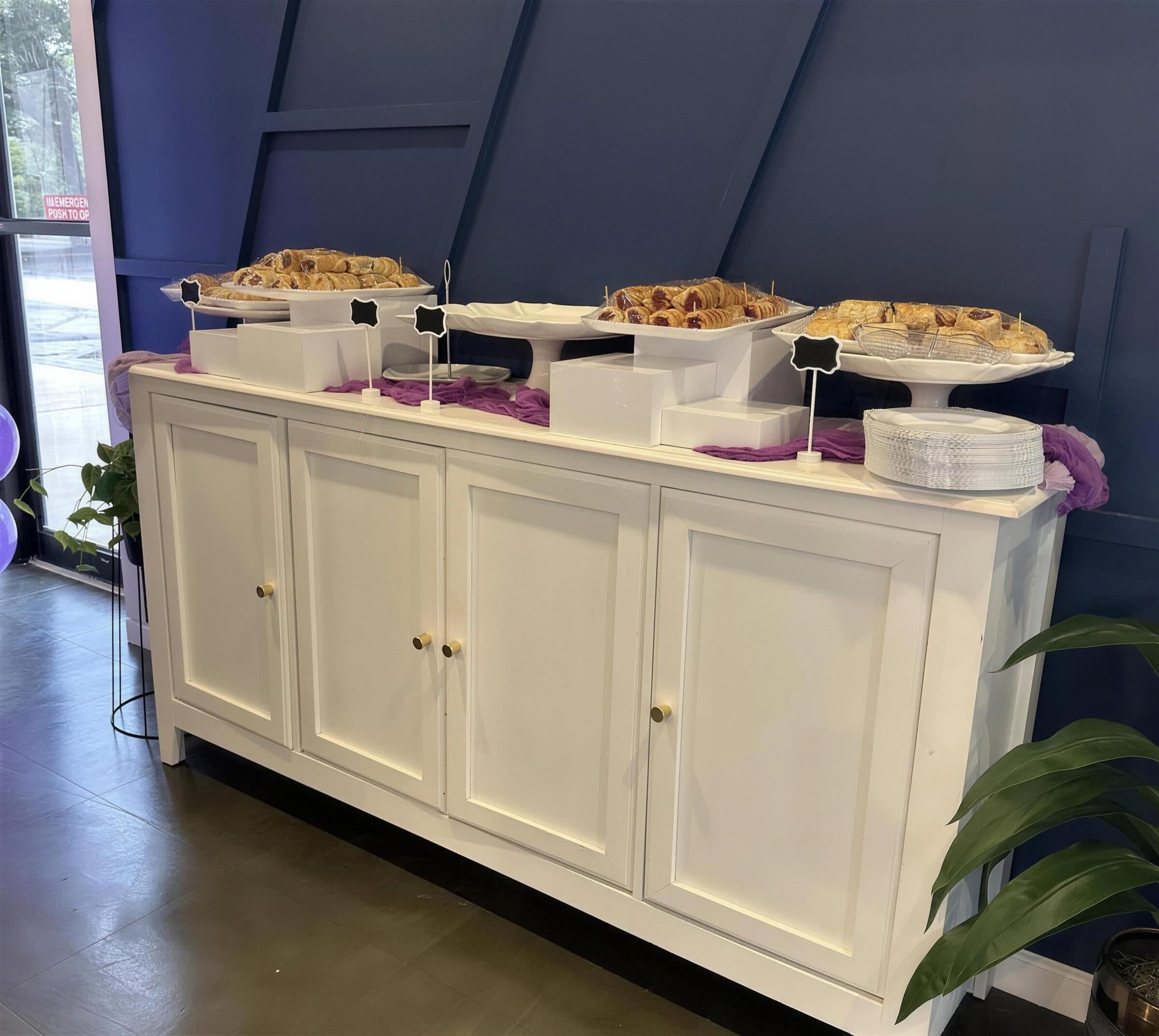 Pastry display on a white sideboard with guava and cheese pastries on cake stands and risers over purple cheesecloth, small chalkboard label tags, at The Cove at St. Johns, Fruit Cove FL