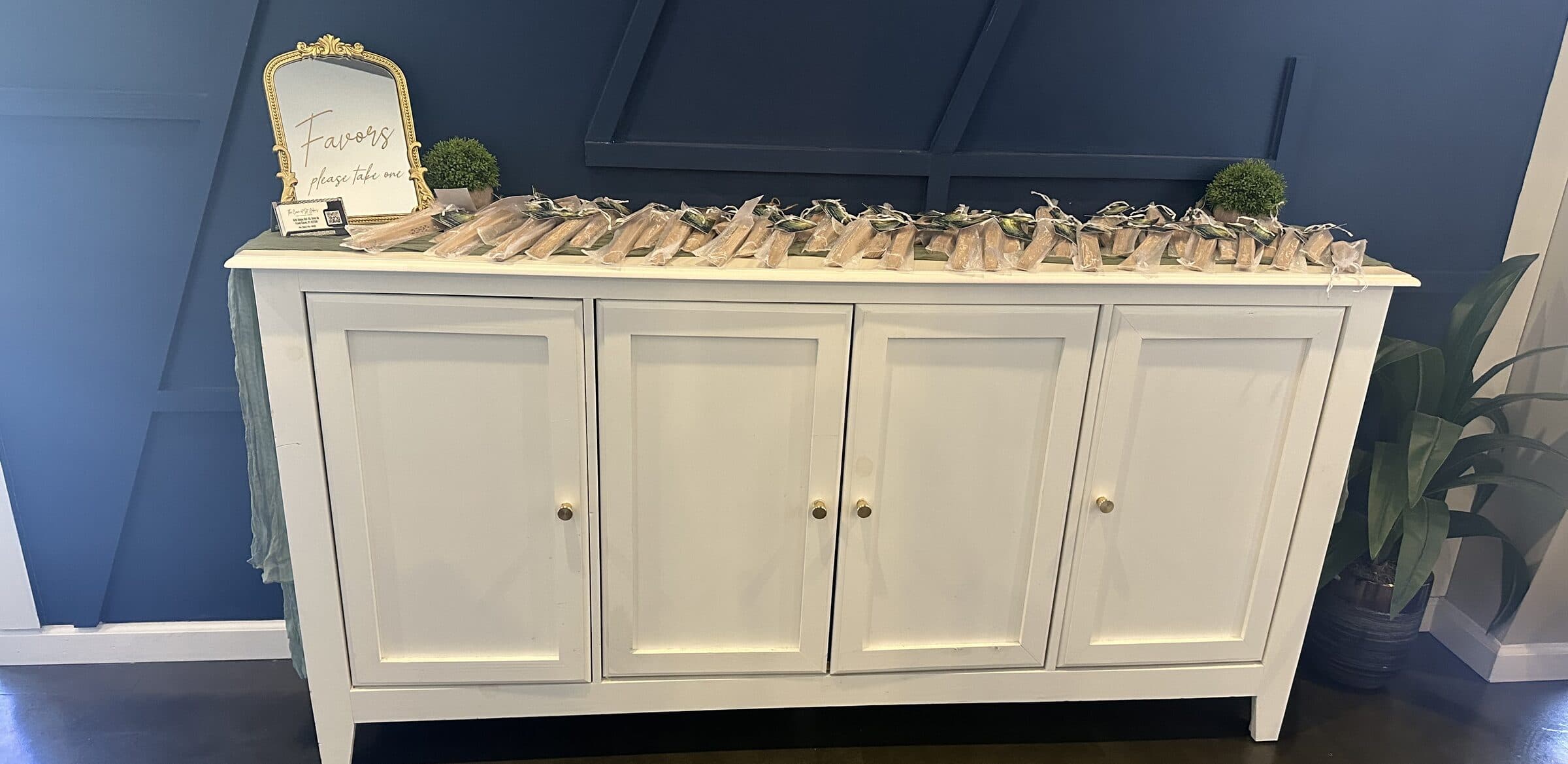 Long row of clear-wrapped treat favors tied with sage ribbon on a white sideboard beside a gold-framed Favors sign and boxwood topiaries at The Cove at St. Johns, near Jacksonville, FL