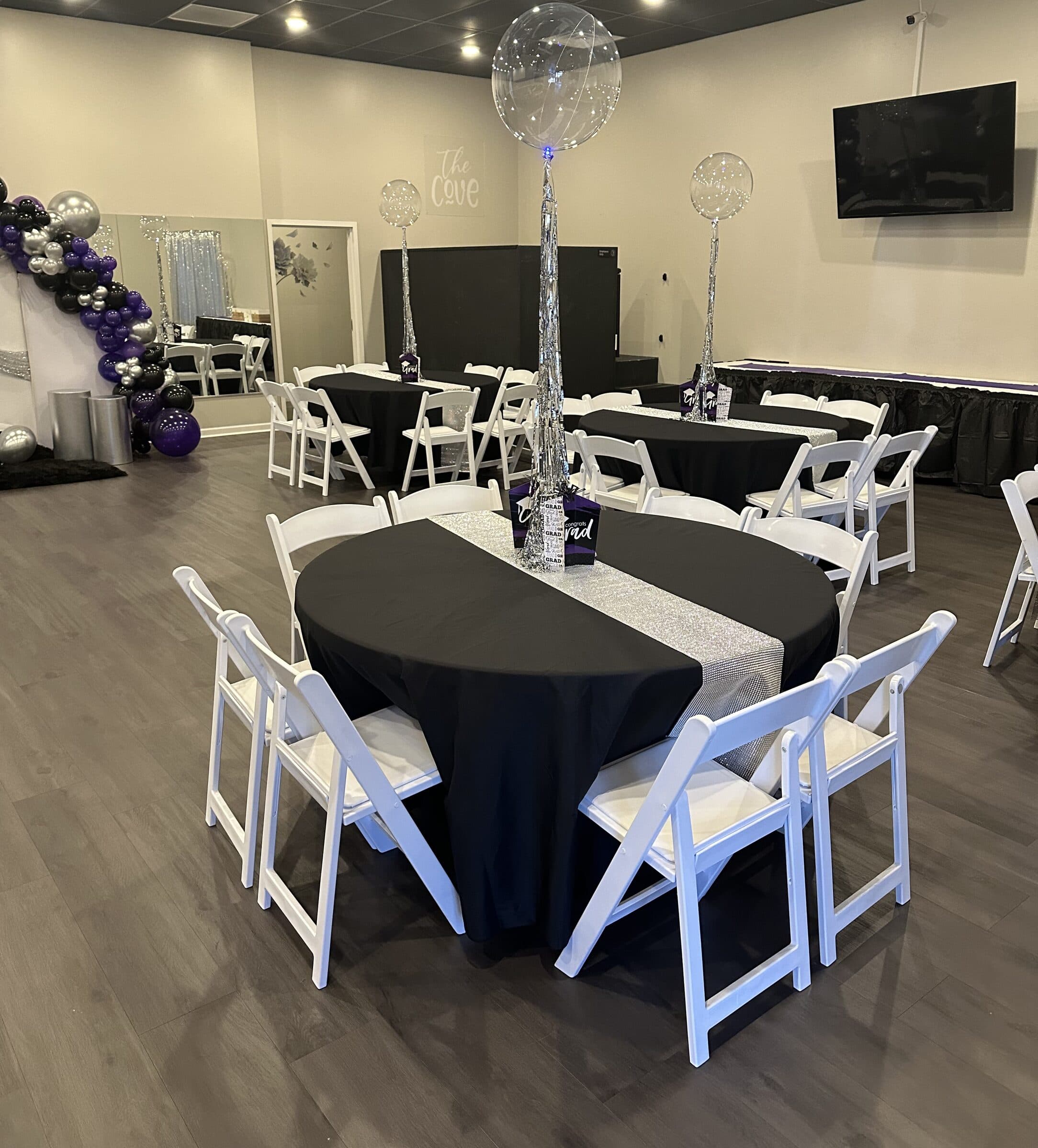 Black-linen round table with a silver sequin runner and Congrats Grad centerpiece under a clear confetti balloon, purple and silver balloon garland behind, at The Cove at St. Johns, Fruit Cove, St. Johns County FL