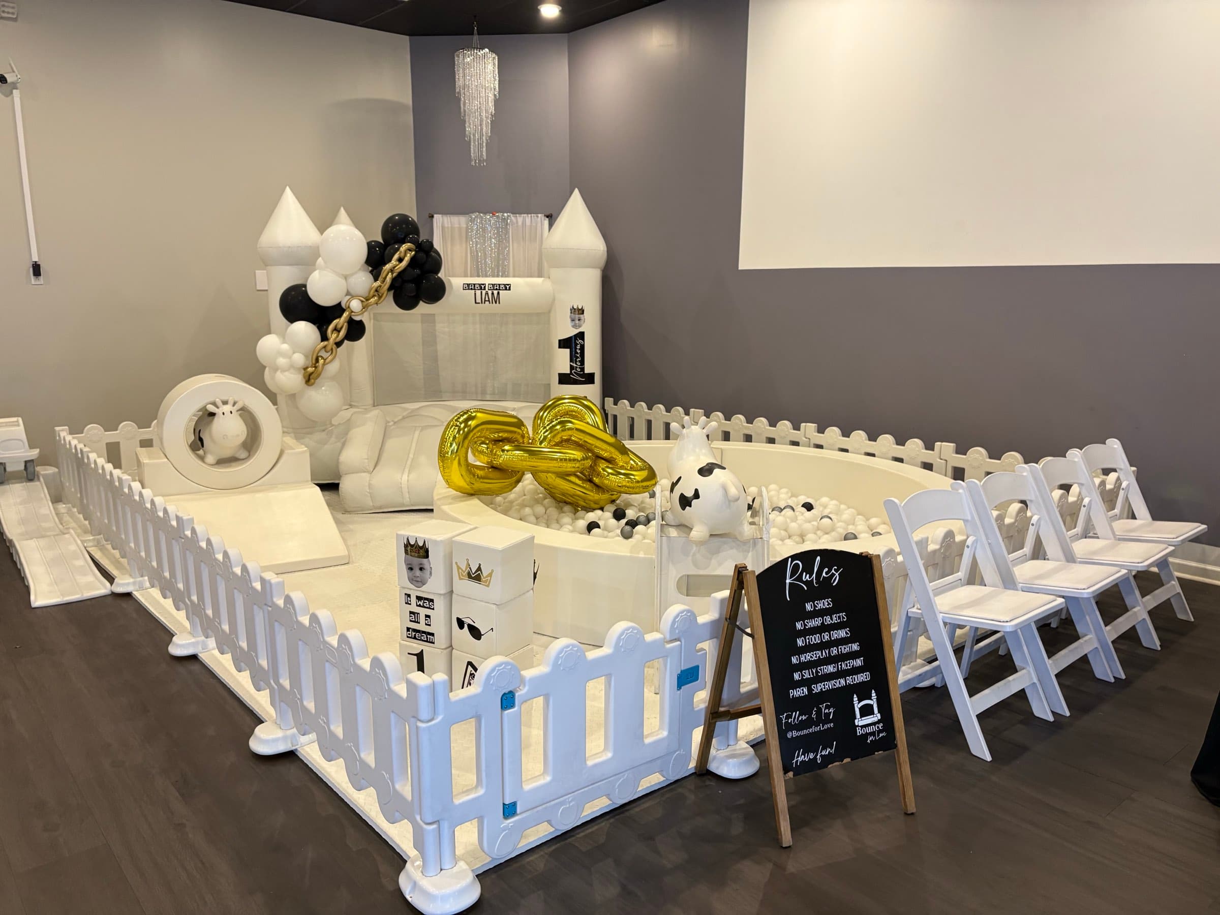 A first birthday party with a white soft-play castle, ball pit and black-and-gold balloon garland at The Cove at St. Johns in Fruit Cove, FL