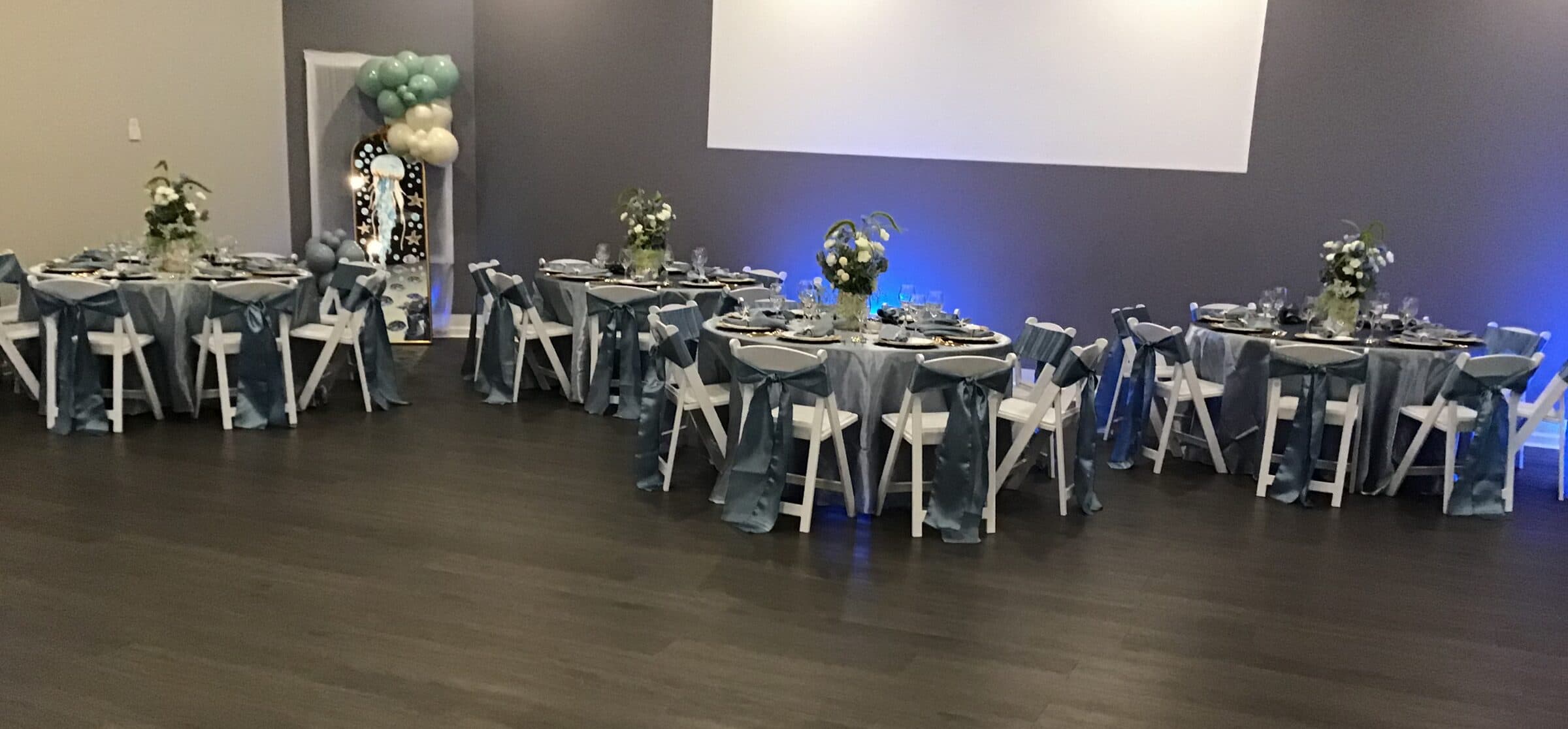 Ground-level angle down steel blue banquet tables with satin chair sashes, white florals and blue uplighting at a birthday party at The Cove at St. Johns, near Jacksonville, FL