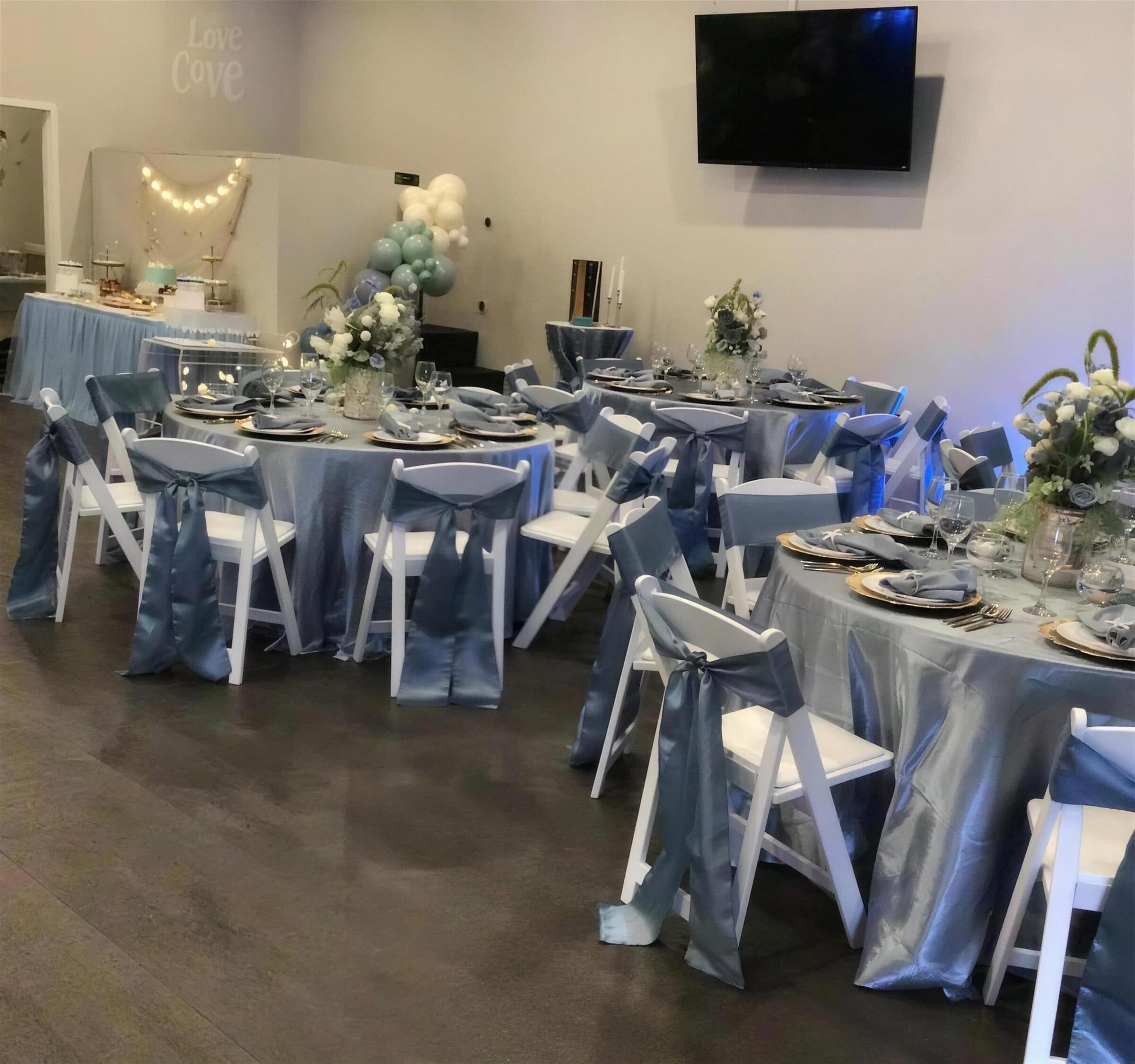 Dessert table with a tiered cake and string lights behind blue satin rounds and sage balloons at a birthday at The Cove at St. Johns, Fruit Cove, St. Johns County FL