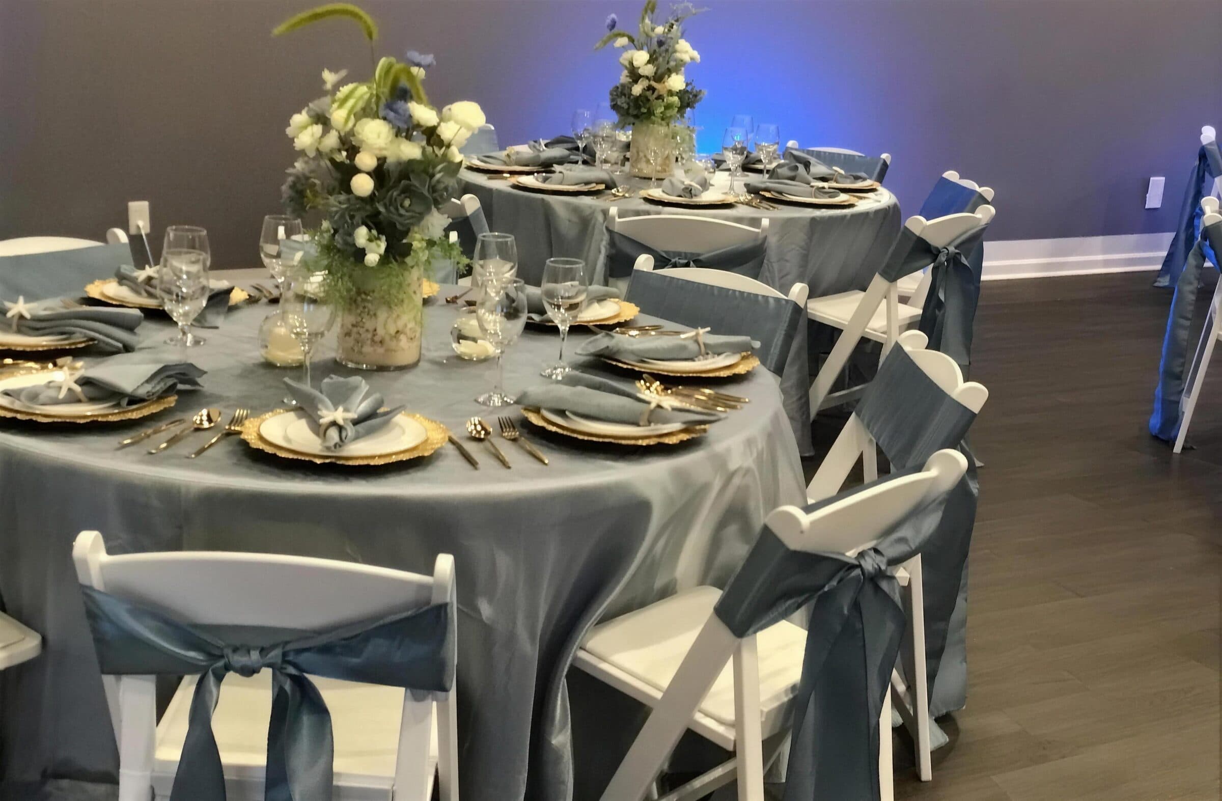 Close-up place setting with gold beaded chargers, blue napkins tied with starfish and a white floral centerpiece at a birthday party at The Cove at St. Johns, Fruit Cove FL