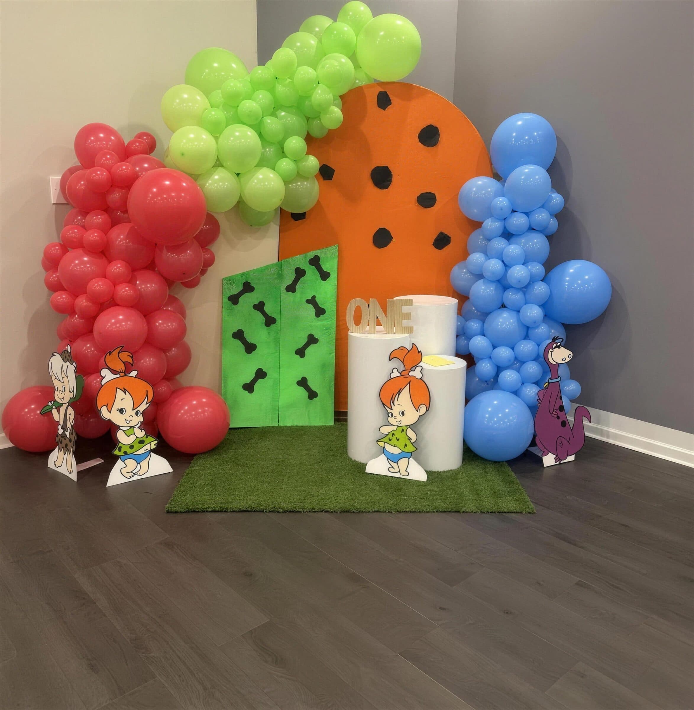 Cartoon-character first birthday backdrop with red, lime and blue balloon columns, an orange arch, faux grass and a wooden ONE sign at The Cove at St. Johns, near Jacksonville, FL