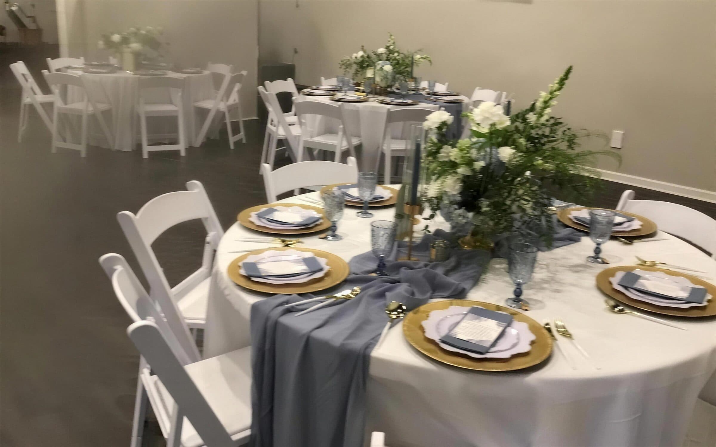 White linens with gold chargers, a slate blue gauze runner, printed menu cards and tall white florals set for a wedding reception at The Cove at St. Johns, Fruit Cove FL