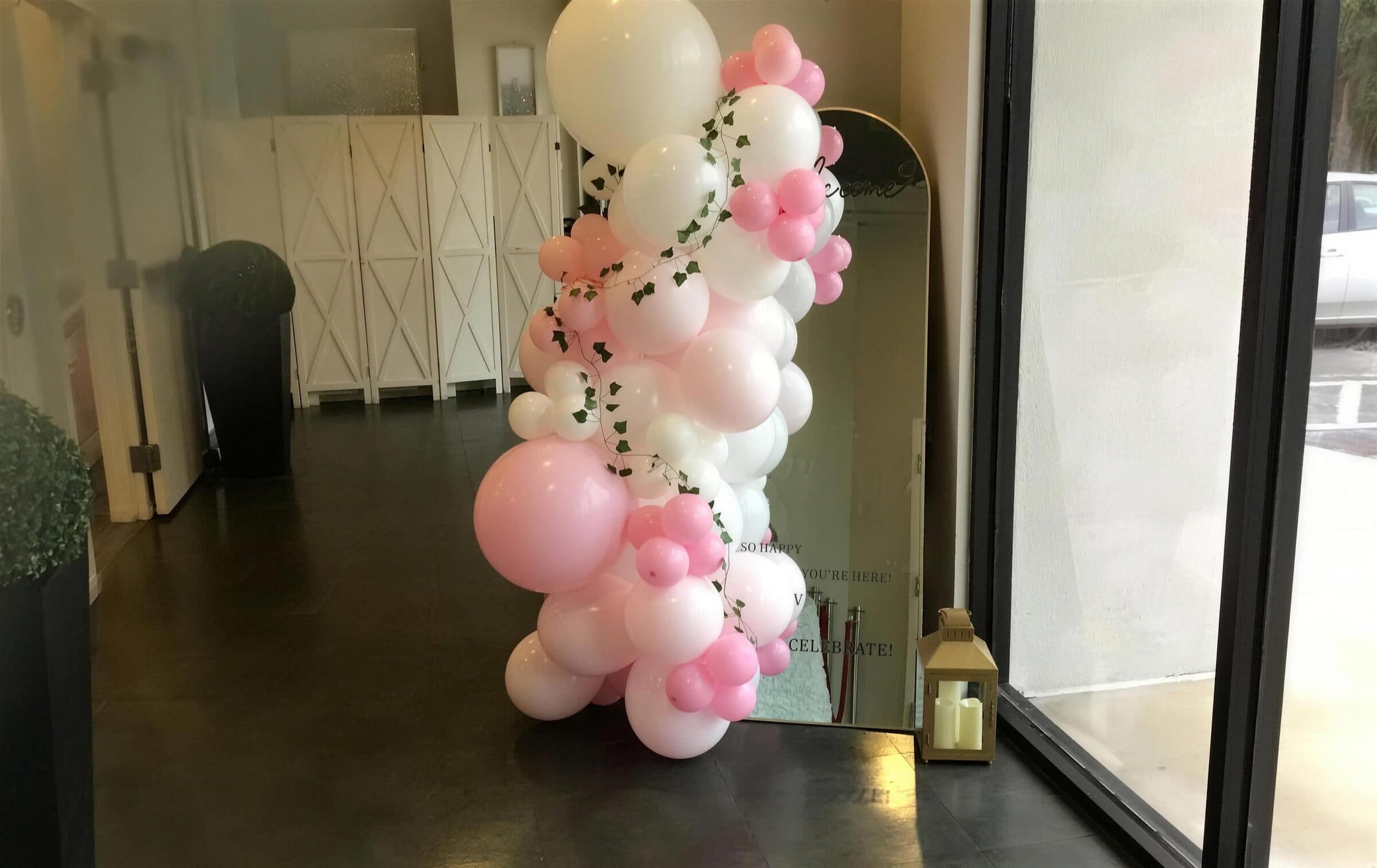 Pink and white balloon garland threaded with ivy framing a gold arched welcome mirror in the sunlit lobby at The Cove at St. Johns, St. Johns County FL