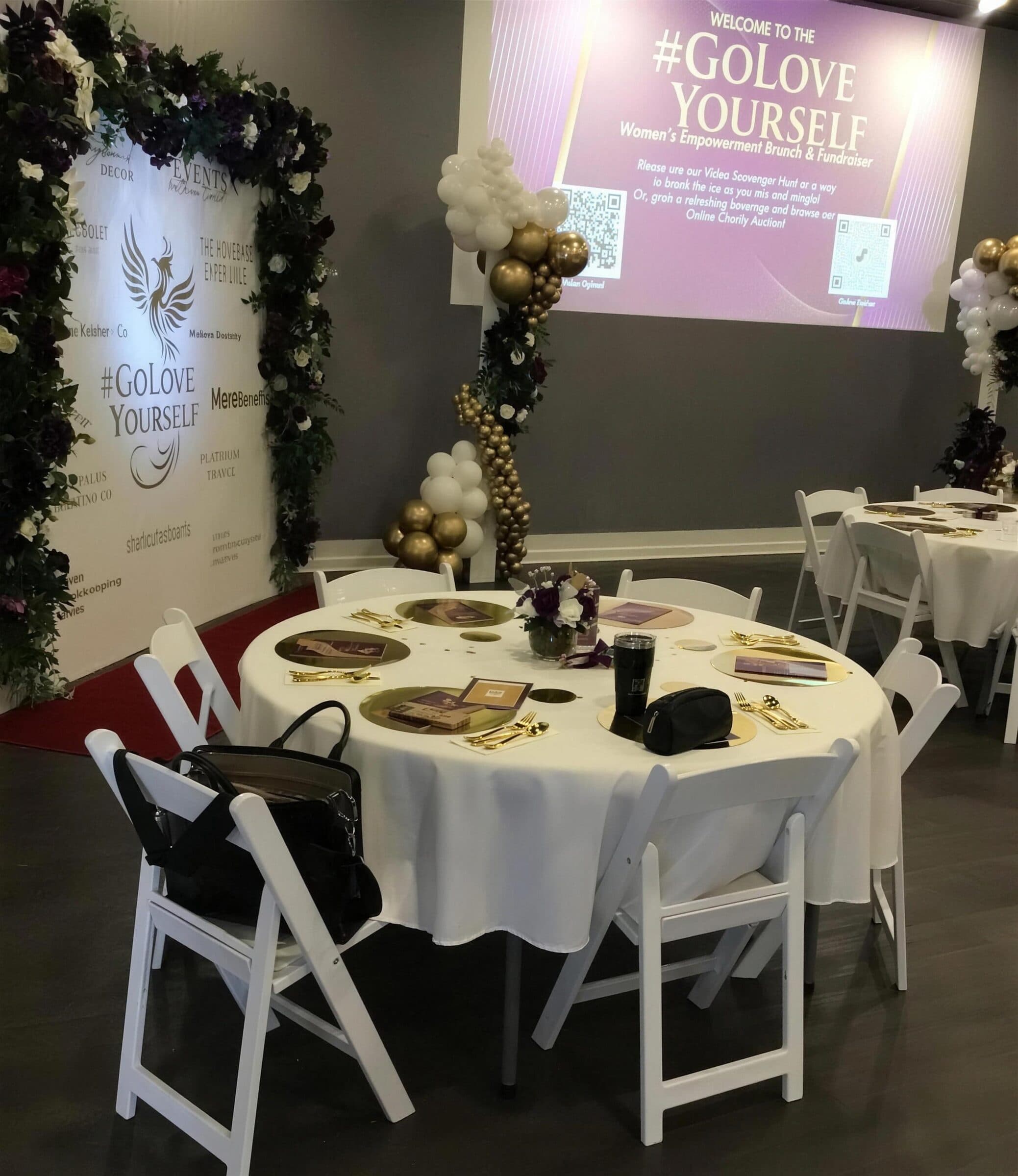 Sponsor step-and-repeat with dark floral garland, gold and white balloon columns and white rounds set for a fundraiser brunch at The Cove at St. Johns, near Jacksonville, FL