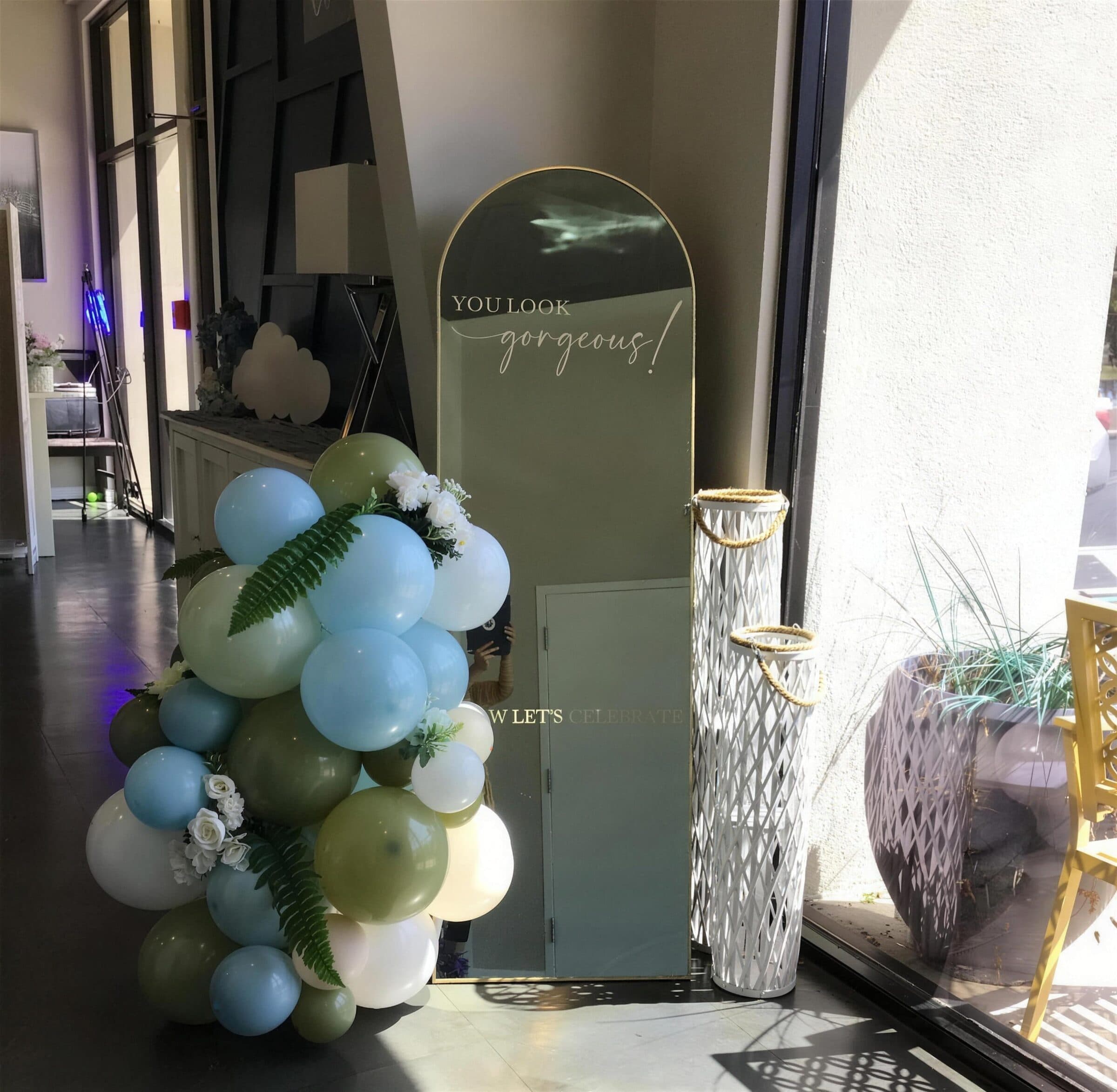 Sage, blue and olive balloon garland with ferns and white blooms beside an arched You Look Gorgeous mirror at The Cove at St. Johns, near Jacksonville, FL