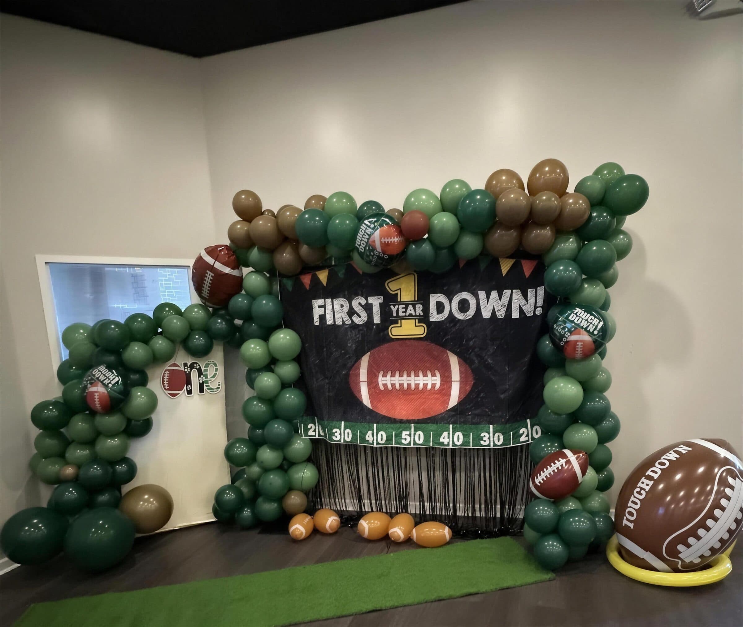 Football first birthday backdrop reading First Year Down with green and brown balloon garland, football foils and a turf runner at The Cove at St. Johns, near Jacksonville, FL