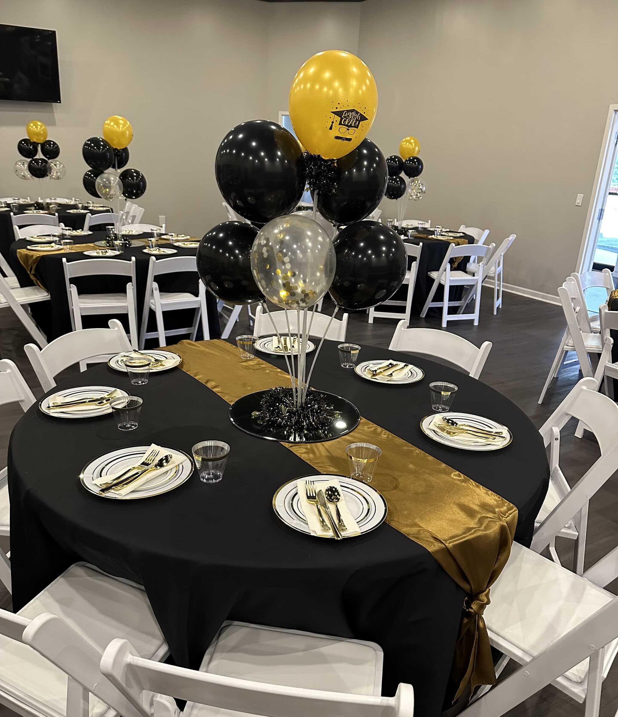 Black and gold graduation table with a gold satin runner, black and confetti balloon centerpiece, white folding chairs and gold flatware at The Cove at St. Johns, St. Johns County FL