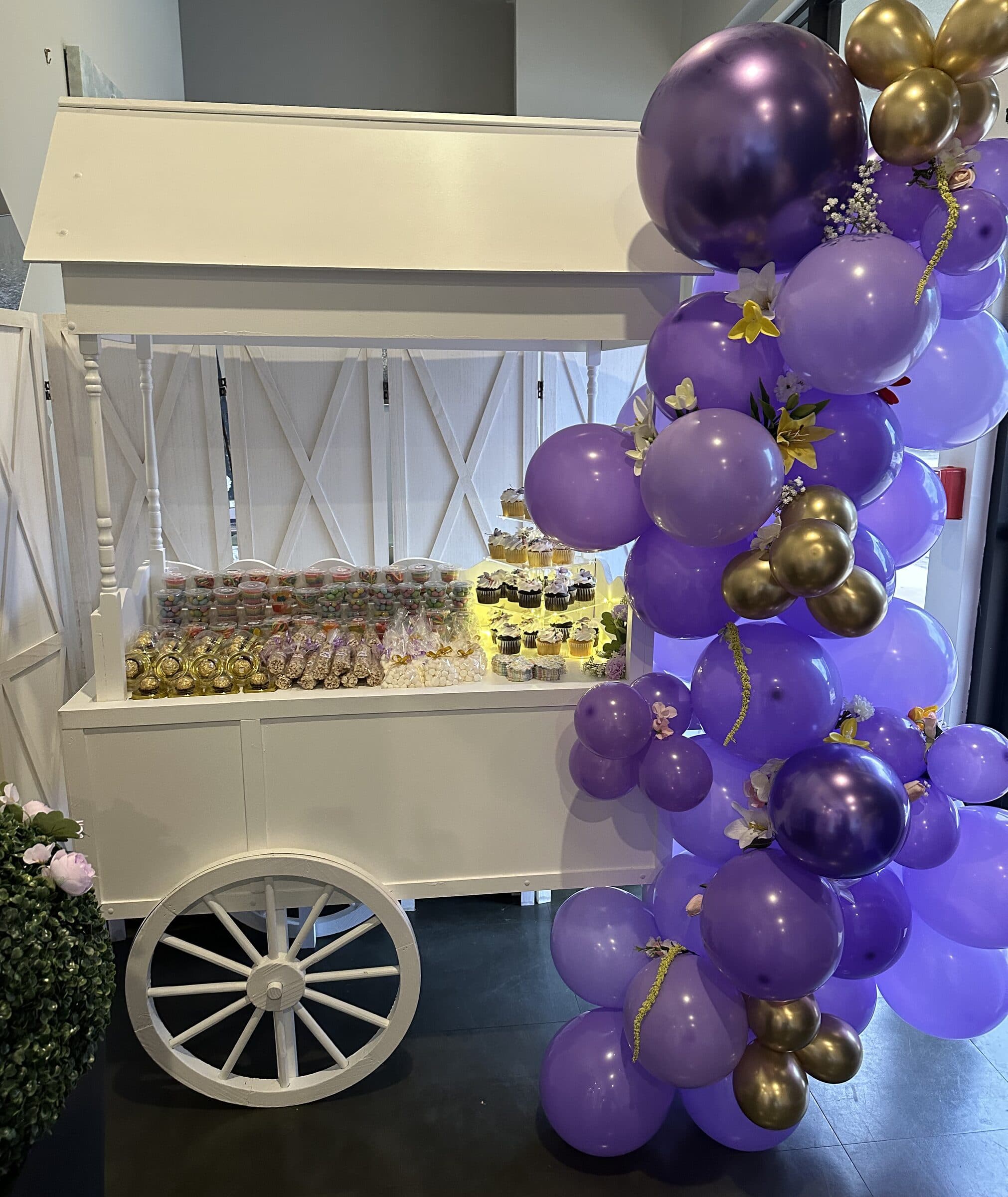 White wooden candy cart loaded with treat jars, cupcakes and favor cones beside a purple and gold balloon garland at a quinceanera at The Cove at St. Johns, near Jacksonville, FL