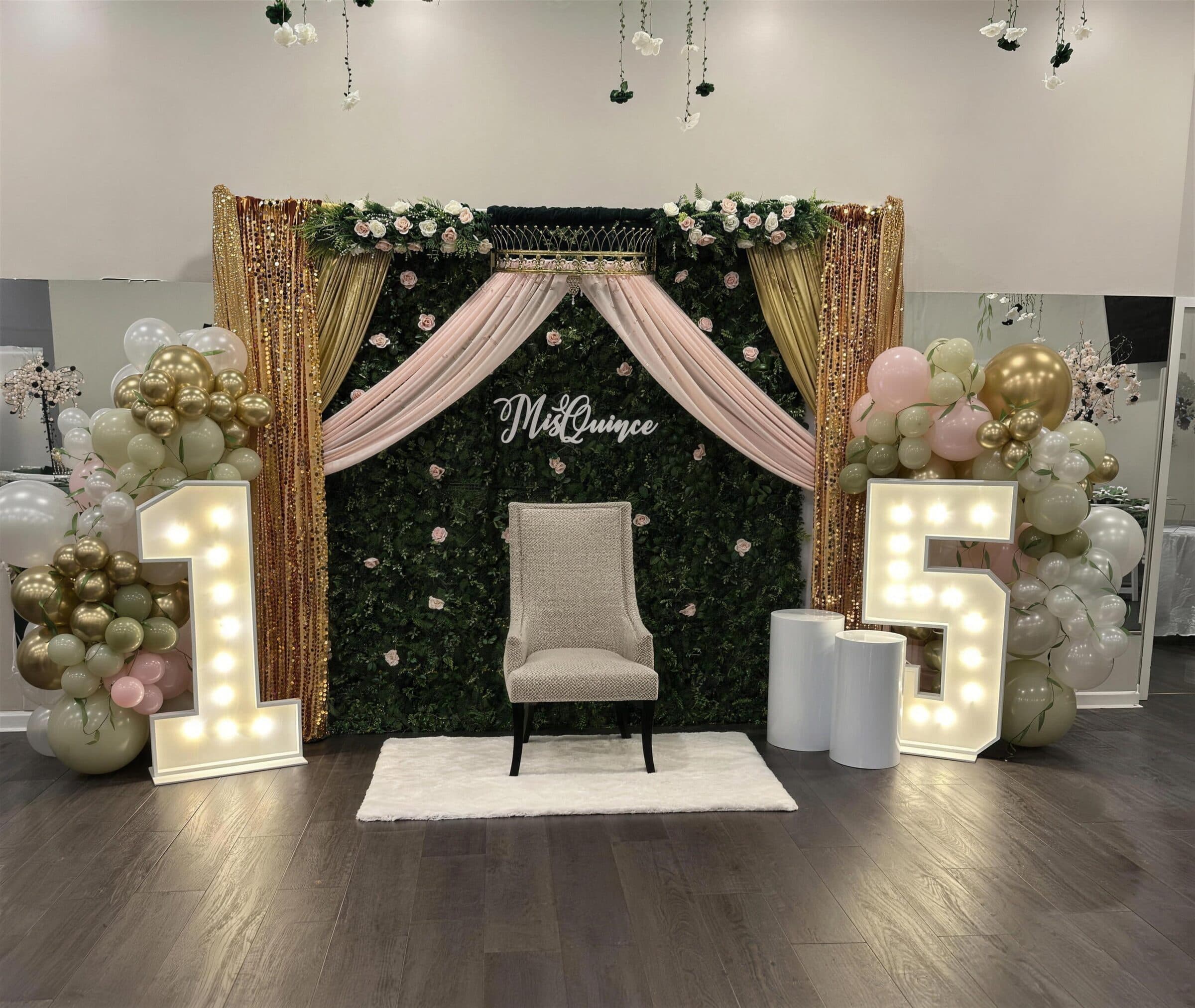 Greenery-wall quinceanera backdrop with blush draping, a gold crown canopy, sage and gold balloons and lit marquee numbers at The Cove at St. Johns, near Jacksonville, FL