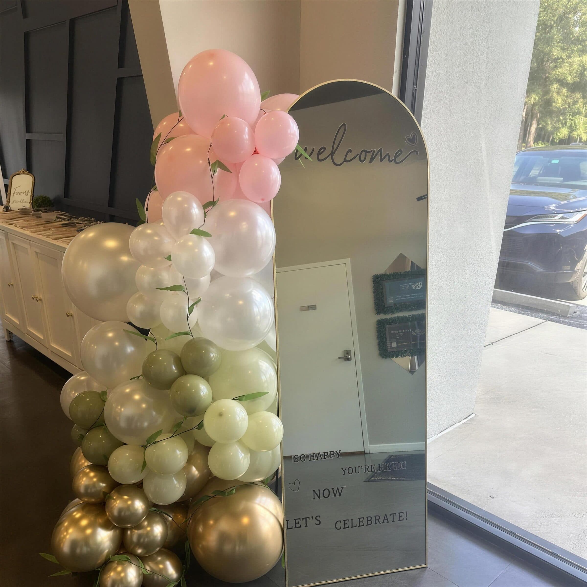 Arched mirror welcome sign reading Let's Celebrate, framed by a blush, ivory, sage and gold balloon garland in the entry at The Cove at St. Johns, Fruit Cove FL