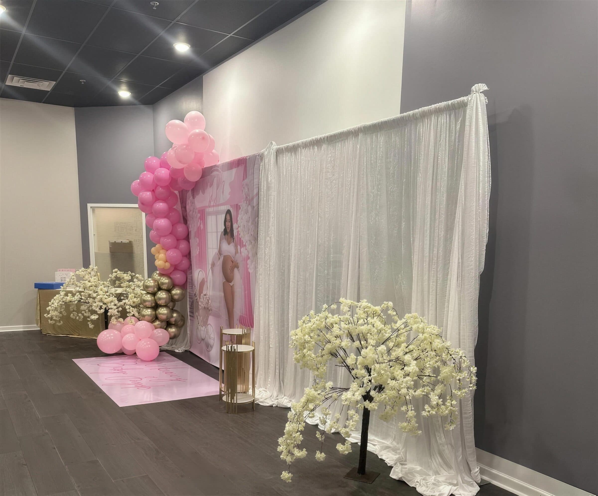 Baby shower photo corner with a pink and gold balloon garland, white shimmer draping, faux blossom trees and a printed pink backdrop at The Cove at St. Johns, near Jacksonville, FL