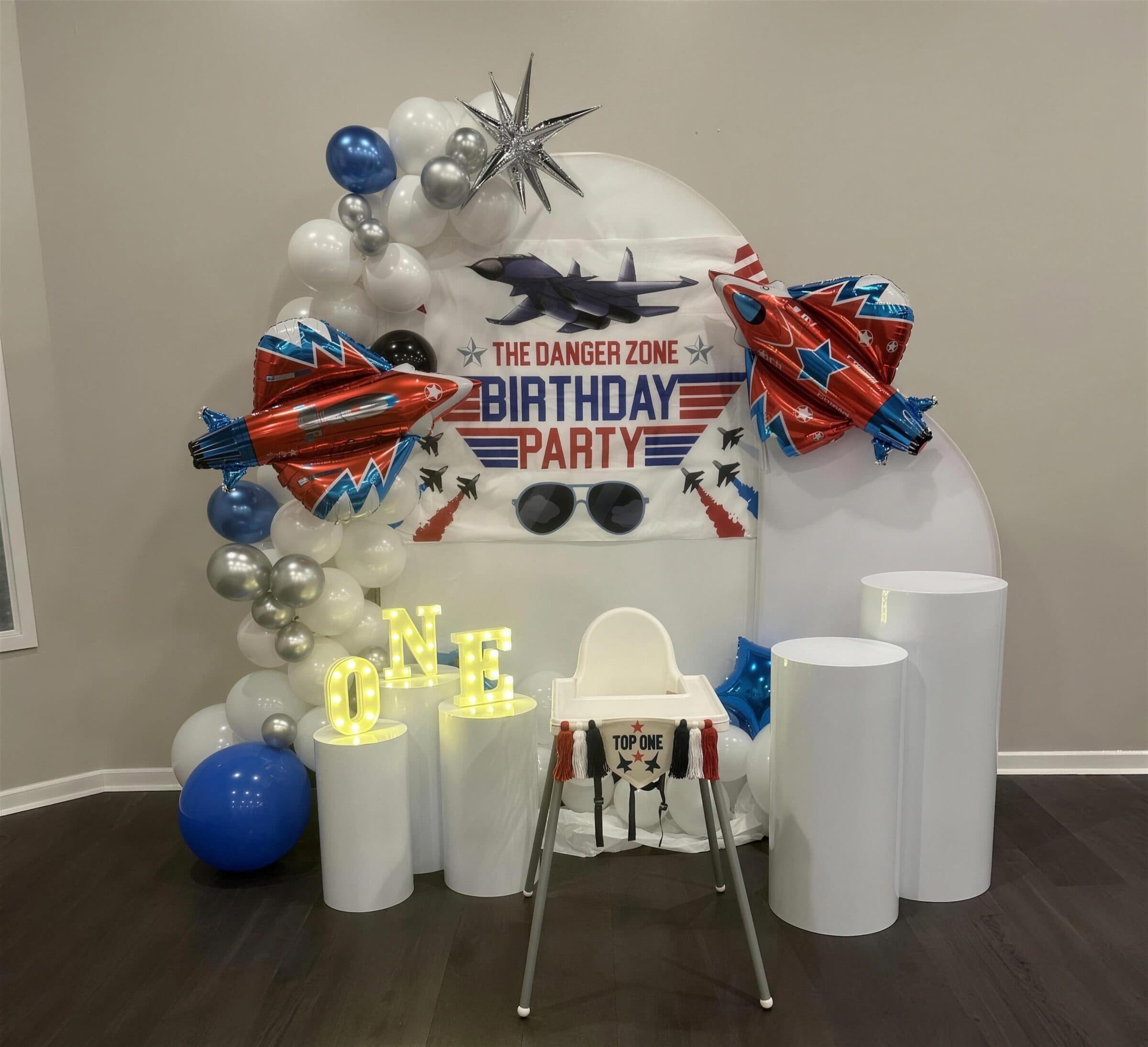 Fighter-jet first birthday setup with red and blue jet balloons, a Danger Zone banner, light-up ONE letters and a decorated high chair at The Cove at St. Johns, Fruit Cove FL