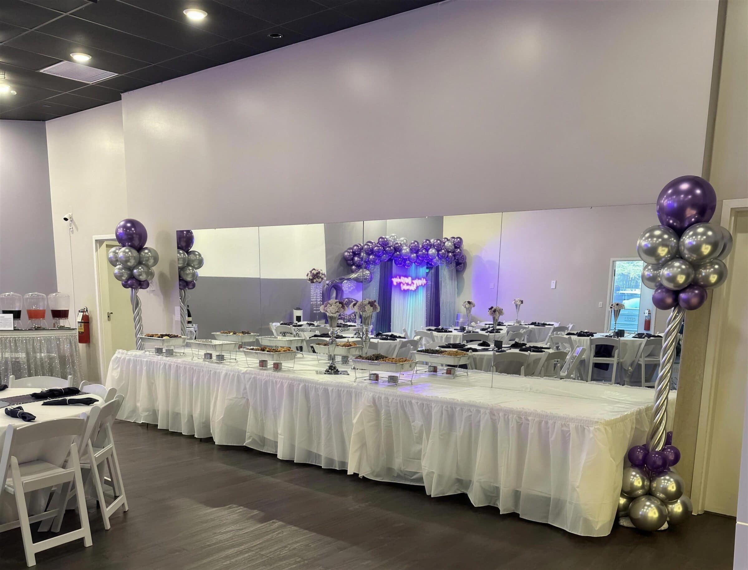 Long skirted buffet line with chafing dishes, floral trumpet vases and purple and silver balloon columns at a 60th birthday at The Cove at St. Johns, near Jacksonville, FL