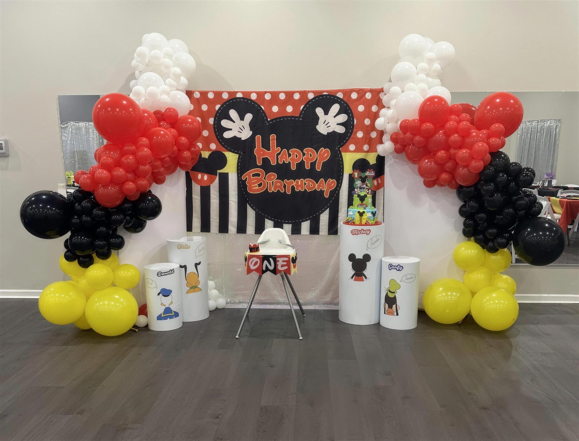 Mickey Mouse first-birthday backdrop with red, black, white and yellow balloon arches, character pedestals and a high chair with a ONE banner at The Cove at St. Johns, Fruit Cove FL
