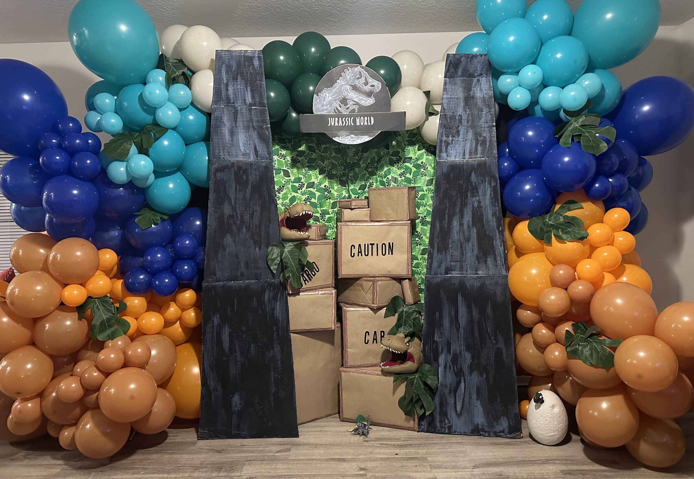 Dinosaur park photo backdrop with teal, blue and orange balloon garlands, painted cardboard gates and stacked CAUTION crates at The Cove at St. Johns, Fruit Cove, St. Johns County FL
