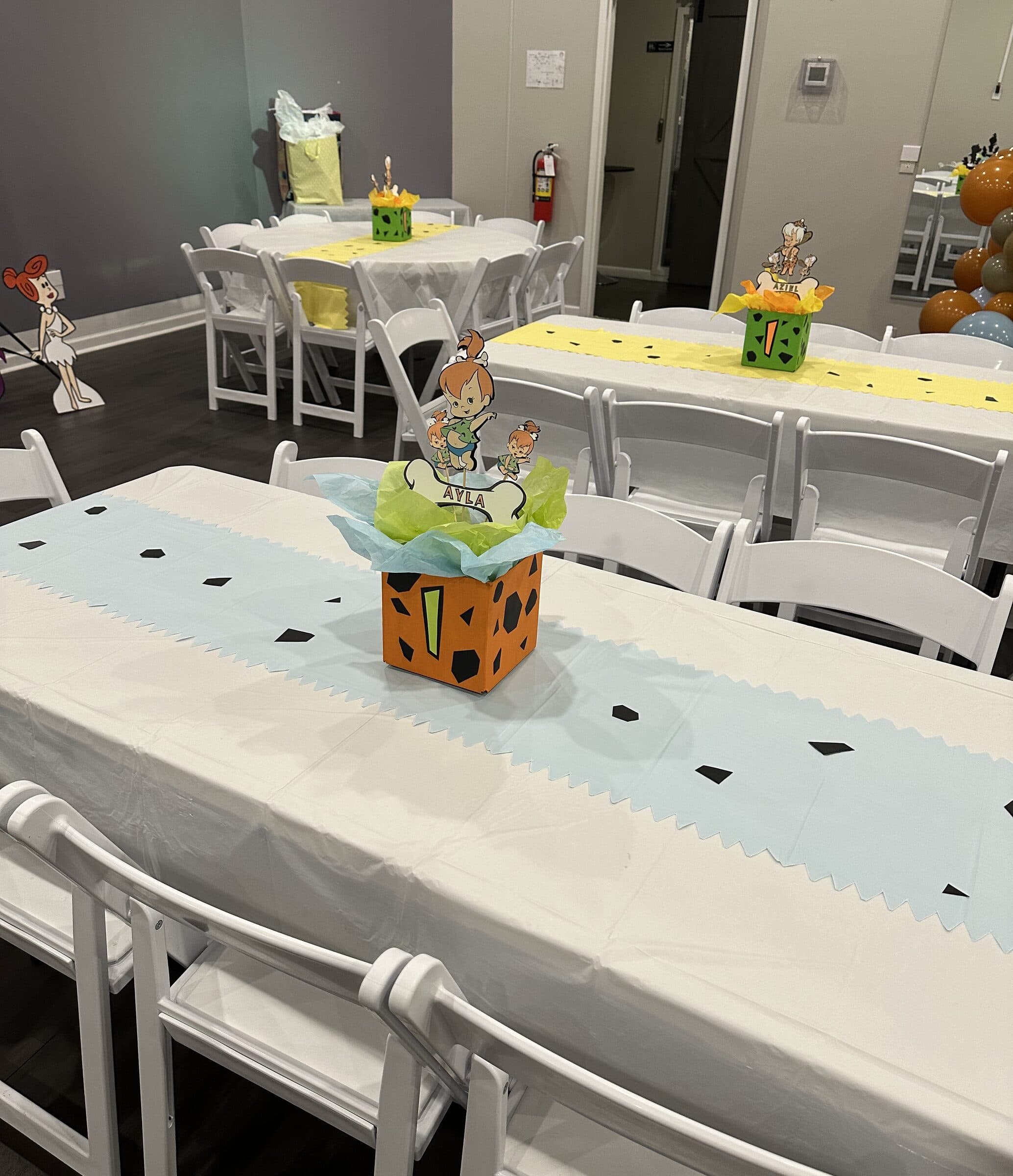 Cartoon cavemen birthday tables at The Cove at St. Johns, near Jacksonville, FL - white linens, blue and yellow zigzag runners and orange stone-print centerpiece boxes with character cutouts