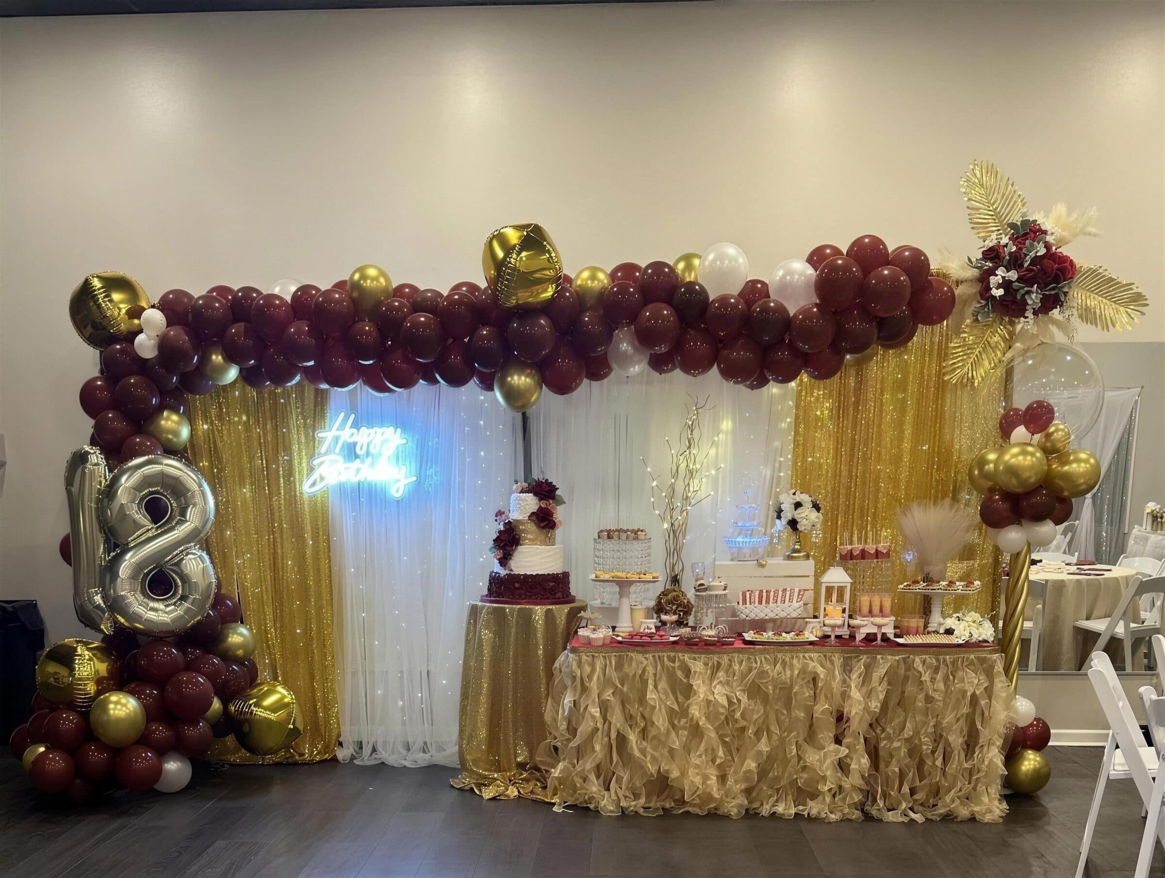 Burgundy and gold 18th birthday setup with balloon garland, gold sequin drapes, Happy Birthday neon, silver number balloons and a tiered cake at The Cove at St. Johns, St. Johns County FL