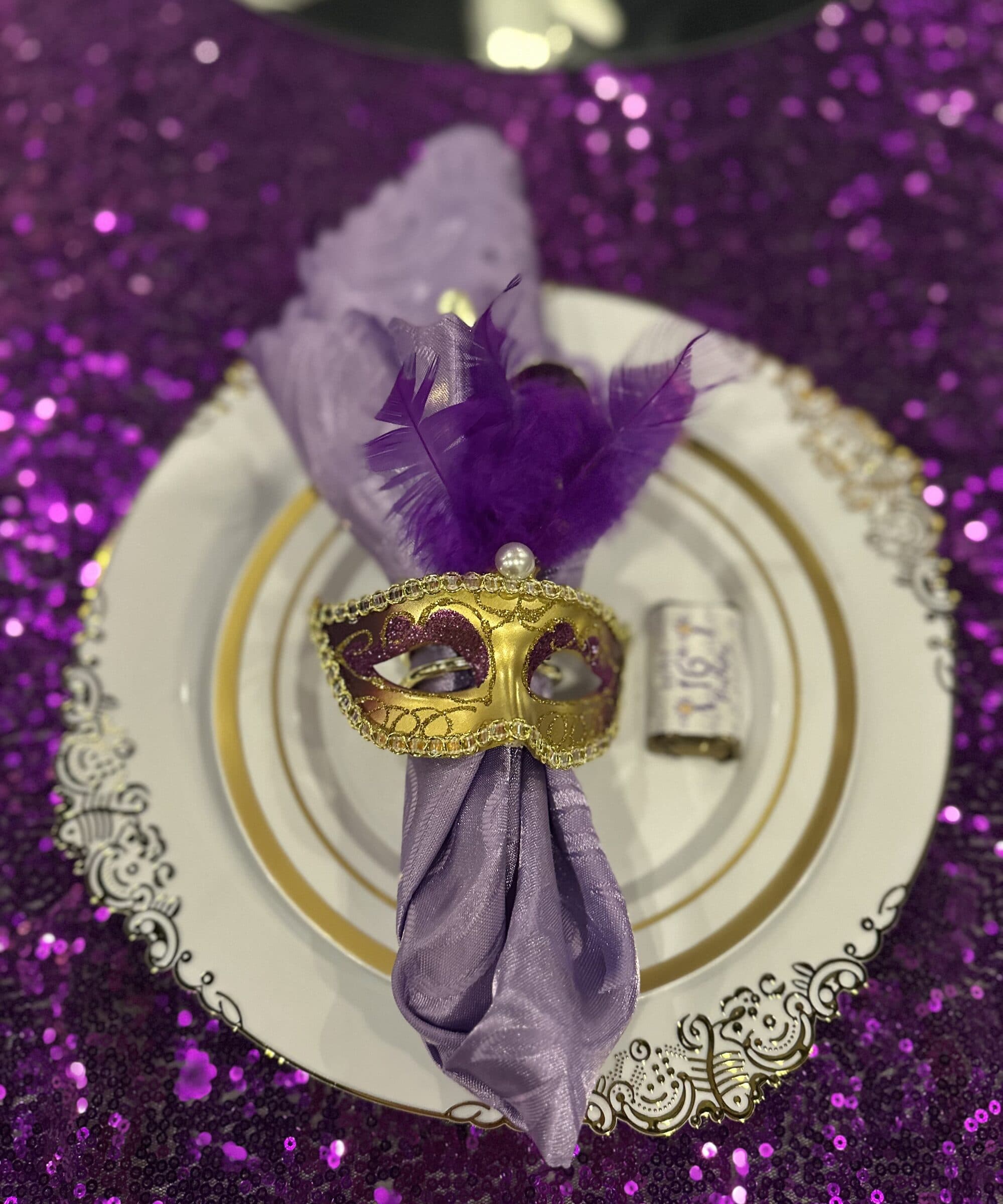 Masquerade place setting close-up at The Cove at St. Johns, Fruit Cove, St. Johns County FL - purple sequin linen, scroll-edge charger and a lilac napkin held by a gold feathered mask