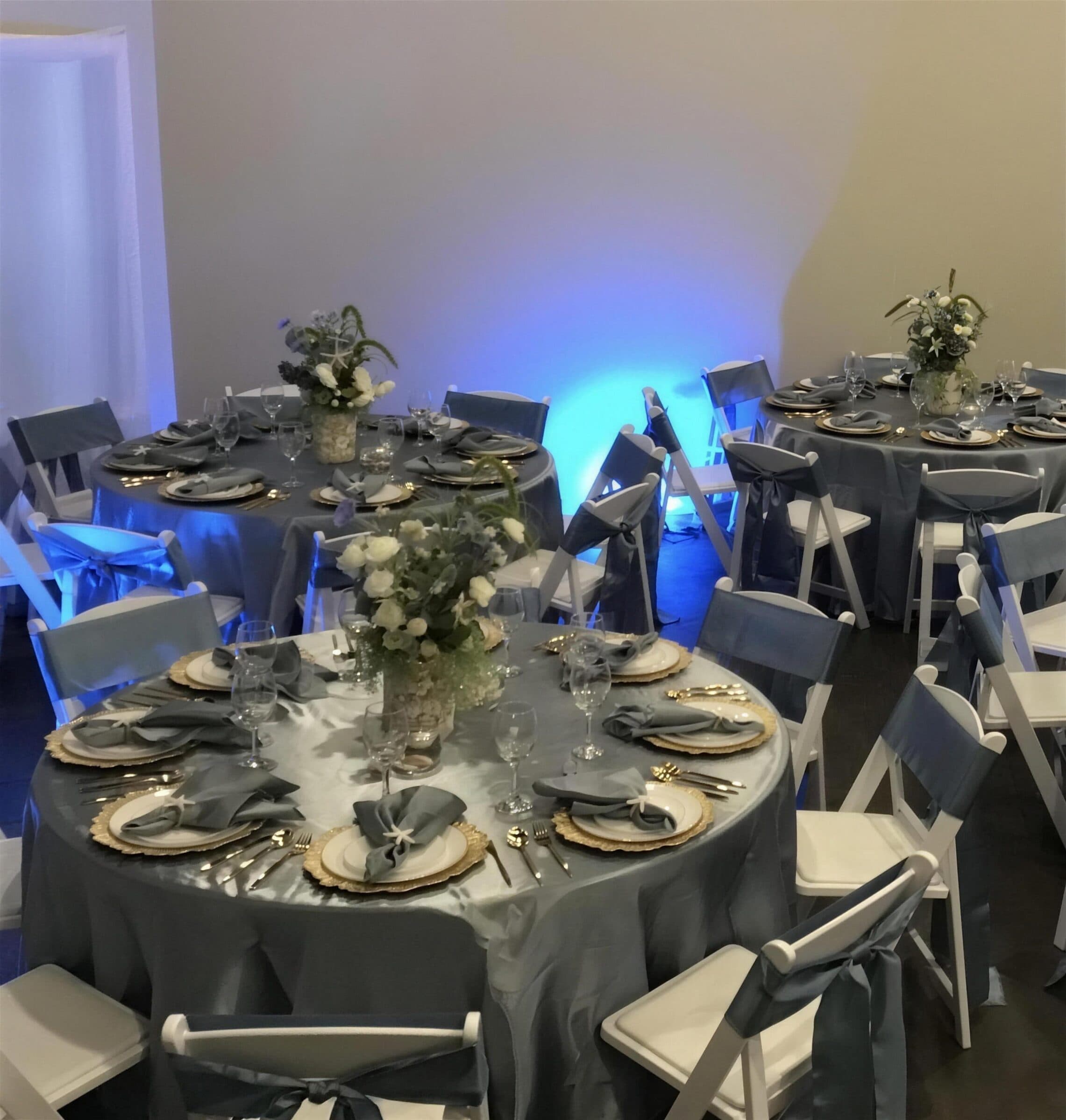 Round tables dressed in slate satin with gold chargers, grey napkins and white garden florals under blue uplighting at The Cove at St. Johns, near Jacksonville, FL