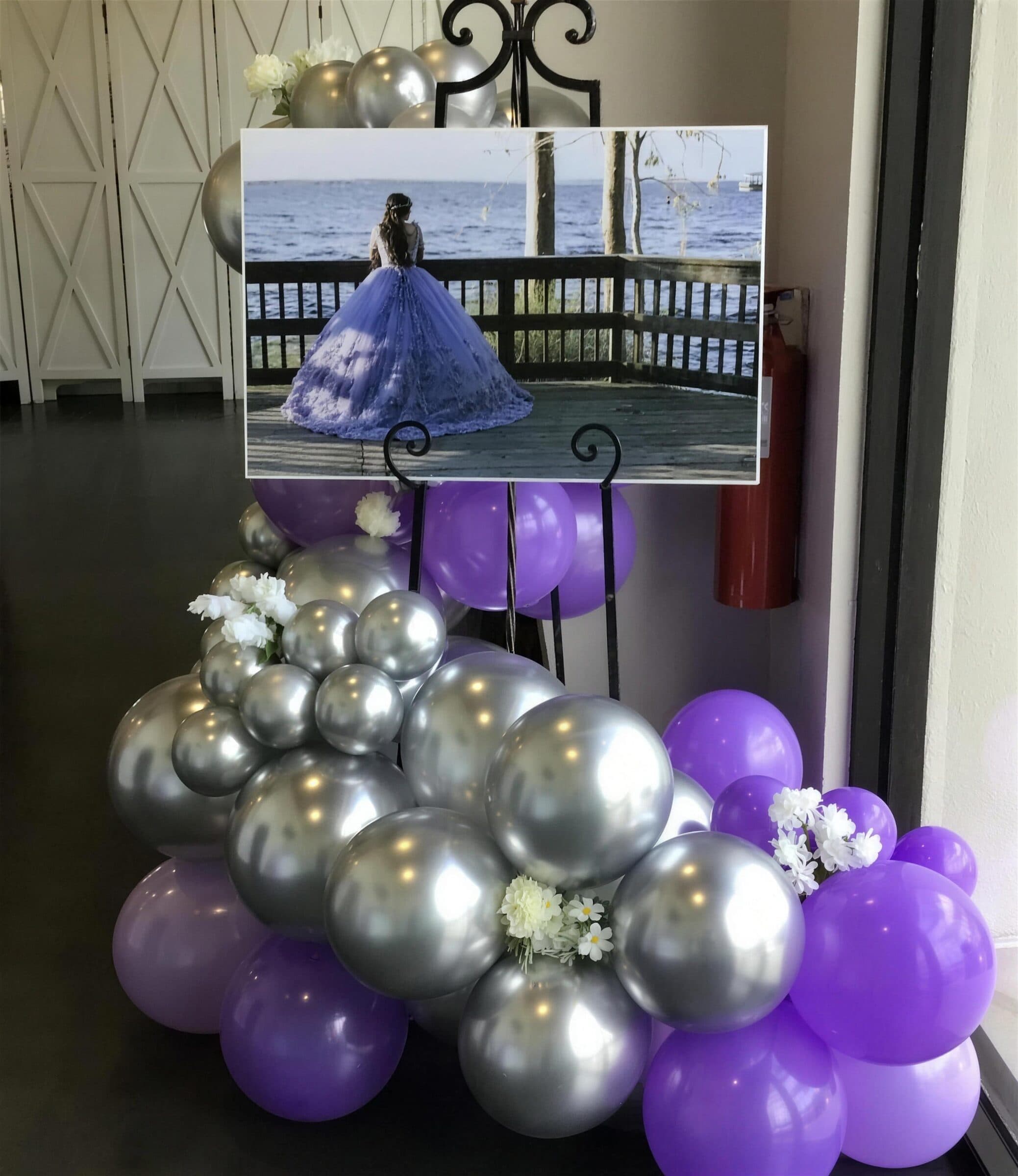 Quinceanera welcome easel holding a large lavender ball-gown portrait, framed by silver chrome and purple balloons with white florals at The Cove at St. Johns, Fruit Cove FL