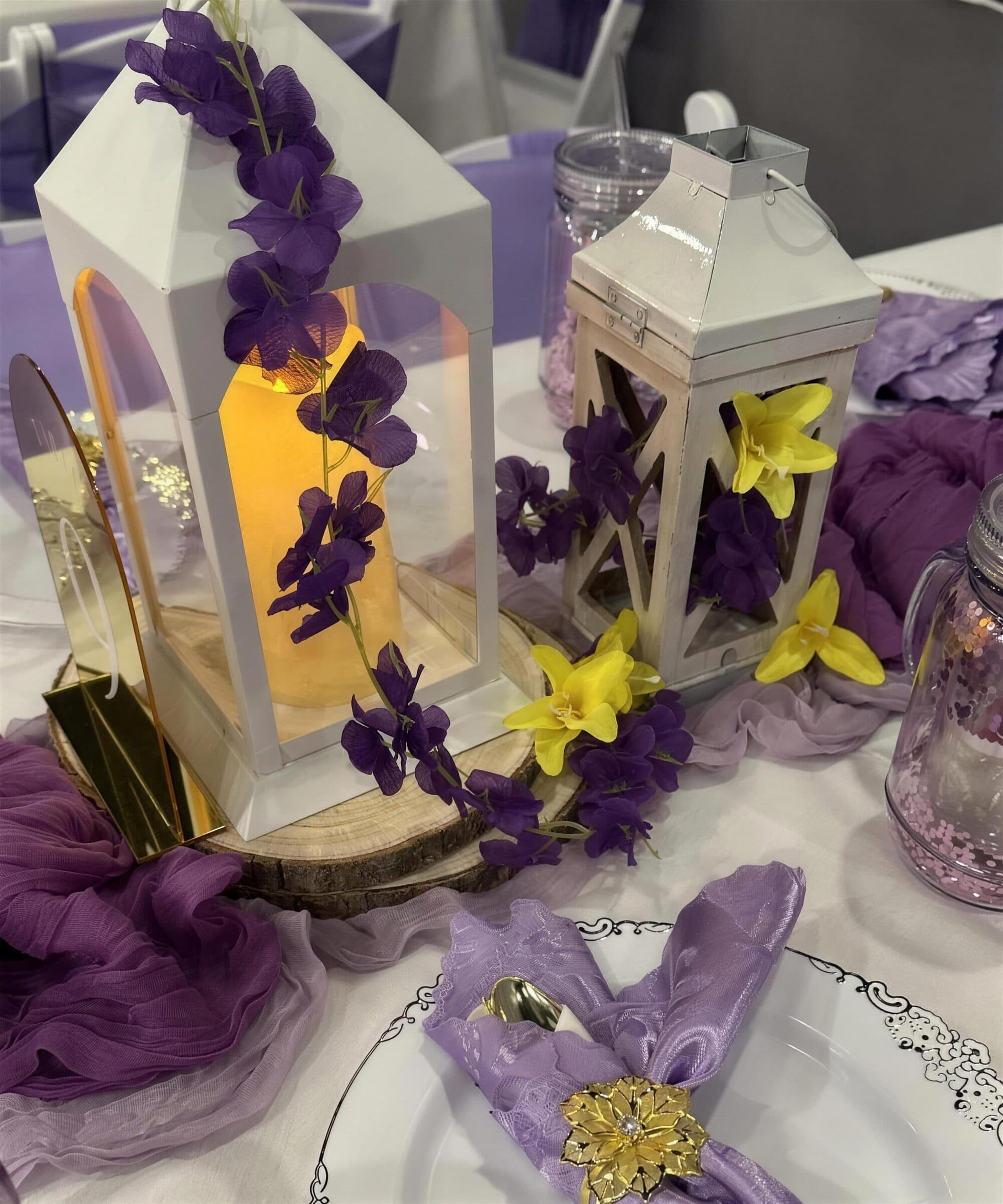 Centerpiece close-up of white lanterns with purple wisteria and yellow blooms on wood slices, lilac chiffon runner and a gold floral napkin ring at The Cove at St. Johns, near Jacksonville, FL
