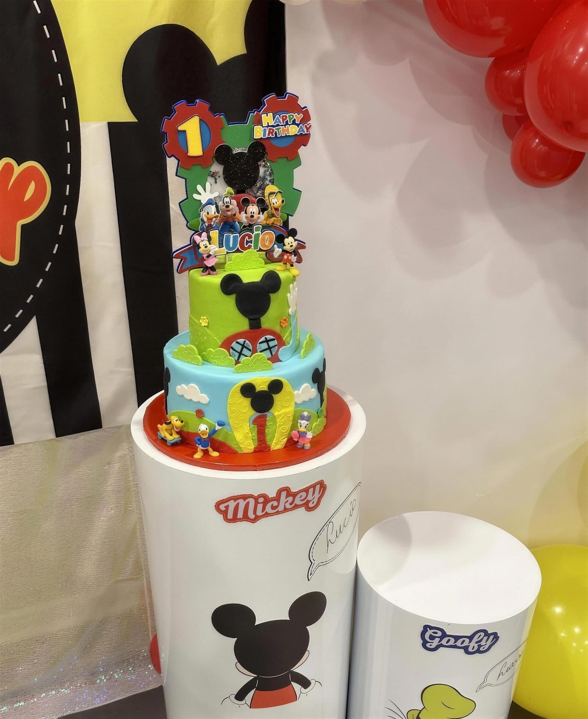 Two-tier green and blue clubhouse cake with fondant figures and a number one topper on a character pedestal beside red balloons at The Cove at St. Johns, Fruit Cove FL