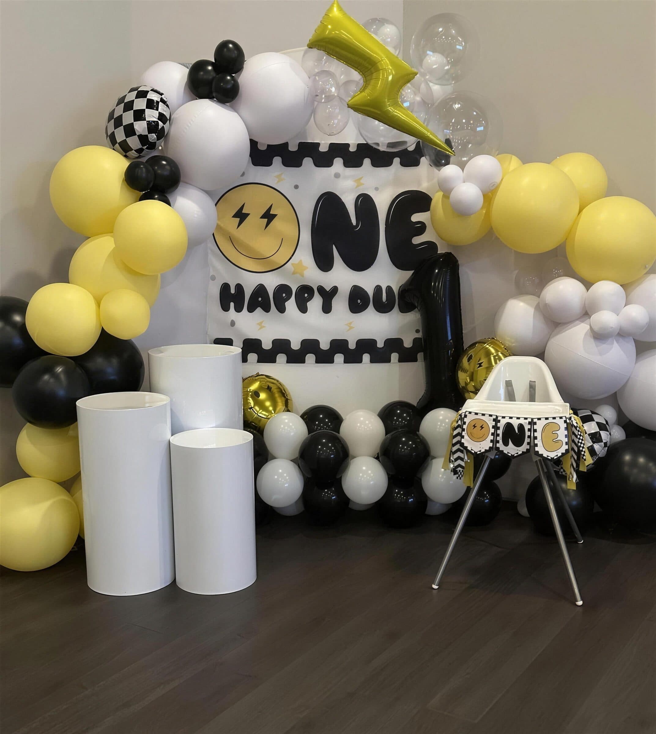 Smiley-face ONE first-birthday backdrop with yellow, black and white balloon garland, checkered and lightning-bolt foils, white pedestals and a banner-trimmed high chair at The Cove at St. Johns, St. Johns County FL