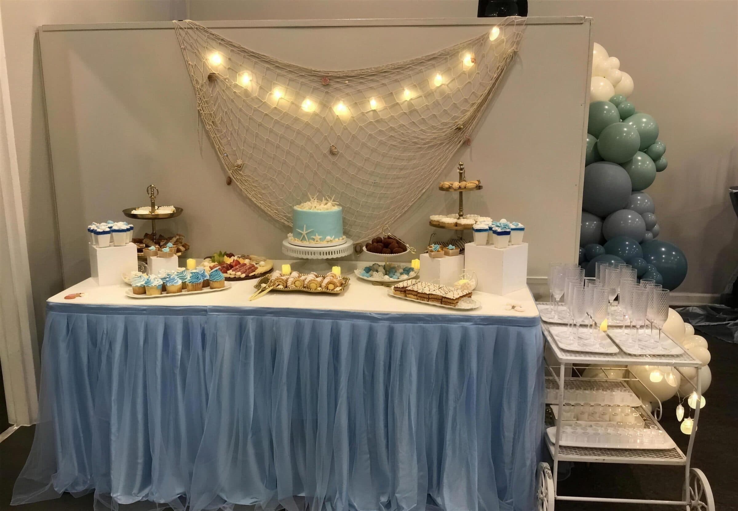 Beach-themed dessert table with a fishing-net and string-light backdrop, pale blue tulle skirt, starfish-topped aqua cake and cupcakes at a baby shower at The Cove at St. Johns, Fruit Cove FL