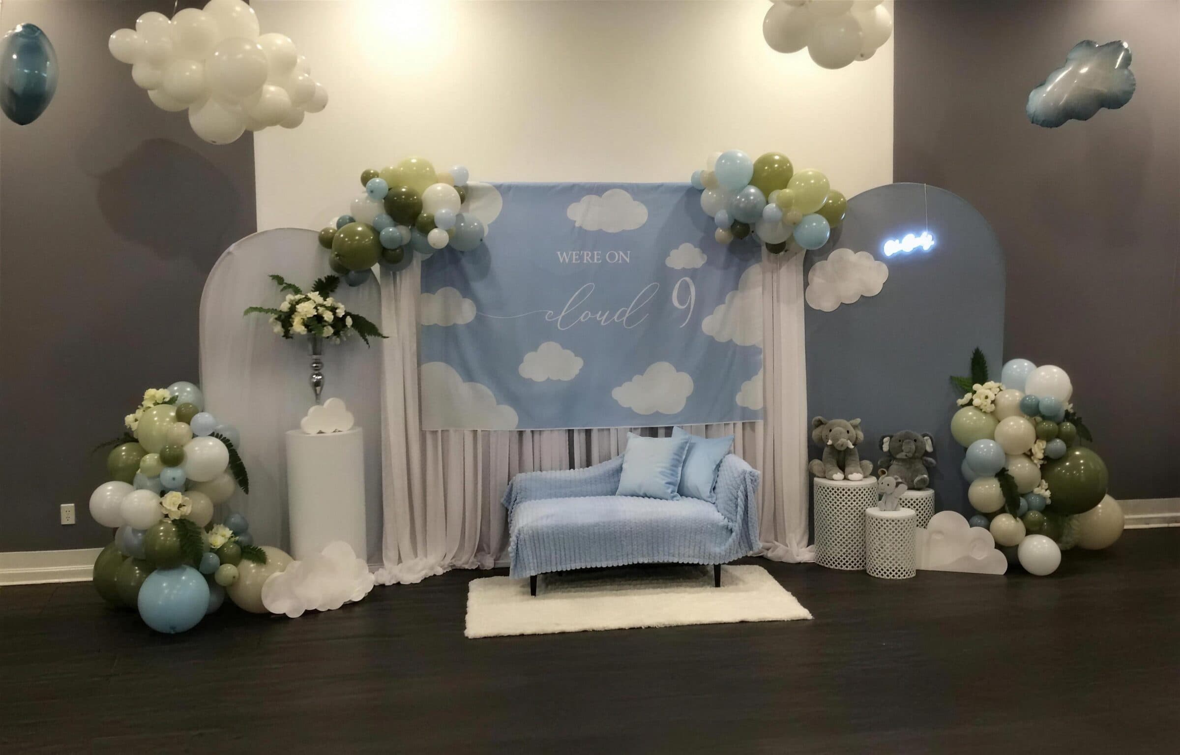 Cloud-nine baby shower photo area with a printed sky backdrop, sage and blue balloon clouds, a blue chaise and elephant plush toys at The Cove at St. Johns, St. Johns County FL