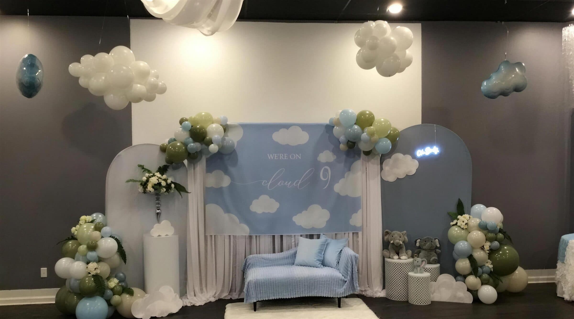 Wider angle on the cloud-themed backdrop, balloon clouds hung from the black ceiling grid above the blue chaise, at a baby shower at The Cove at St. Johns, near Jacksonville, FL