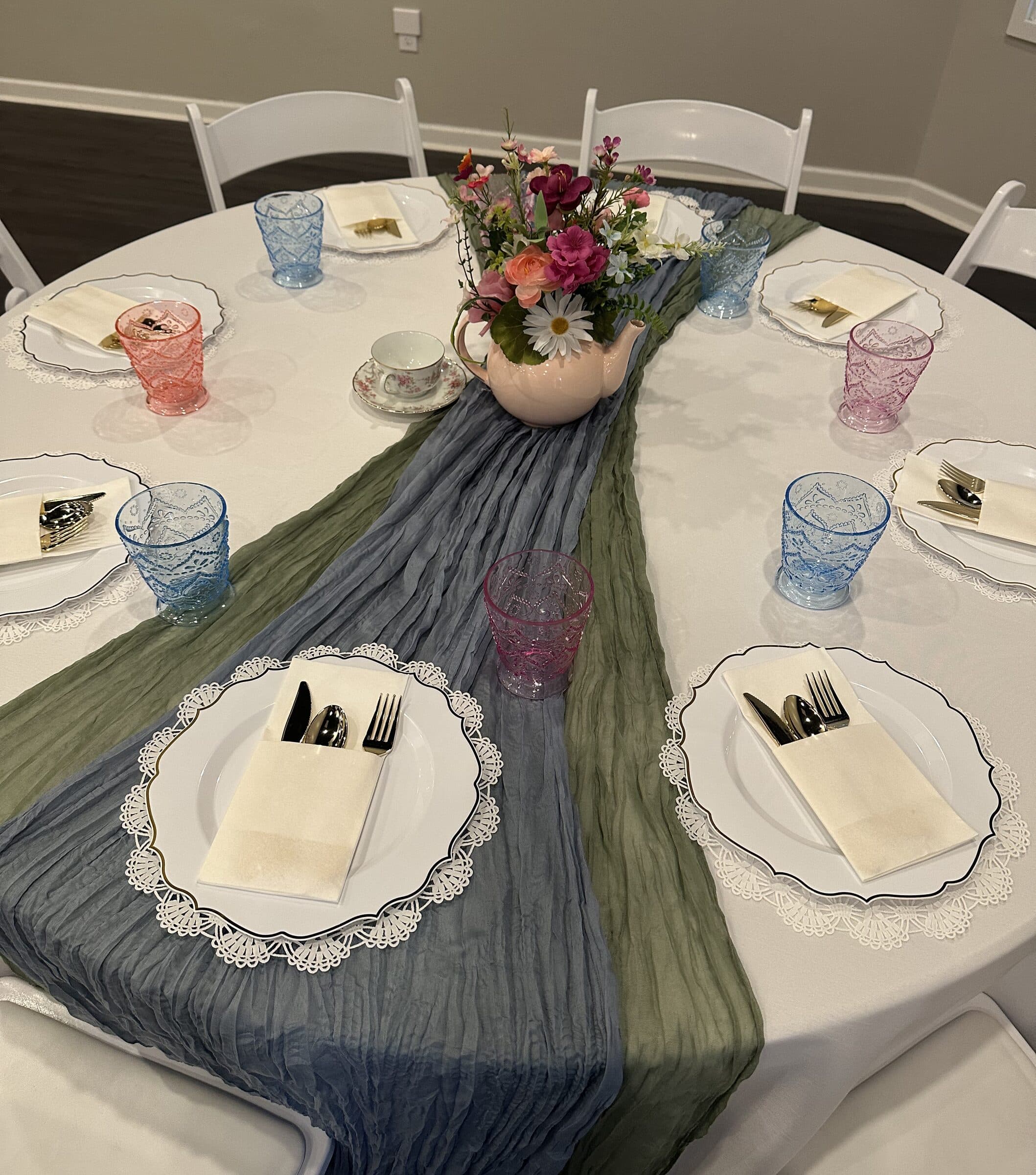 Bridal tea table set with white lace-edged chargers, blue and sage cheesecloth runners, colored pressed-glass goblets and a floral teapot at The Cove at St. Johns, near Jacksonville, FL