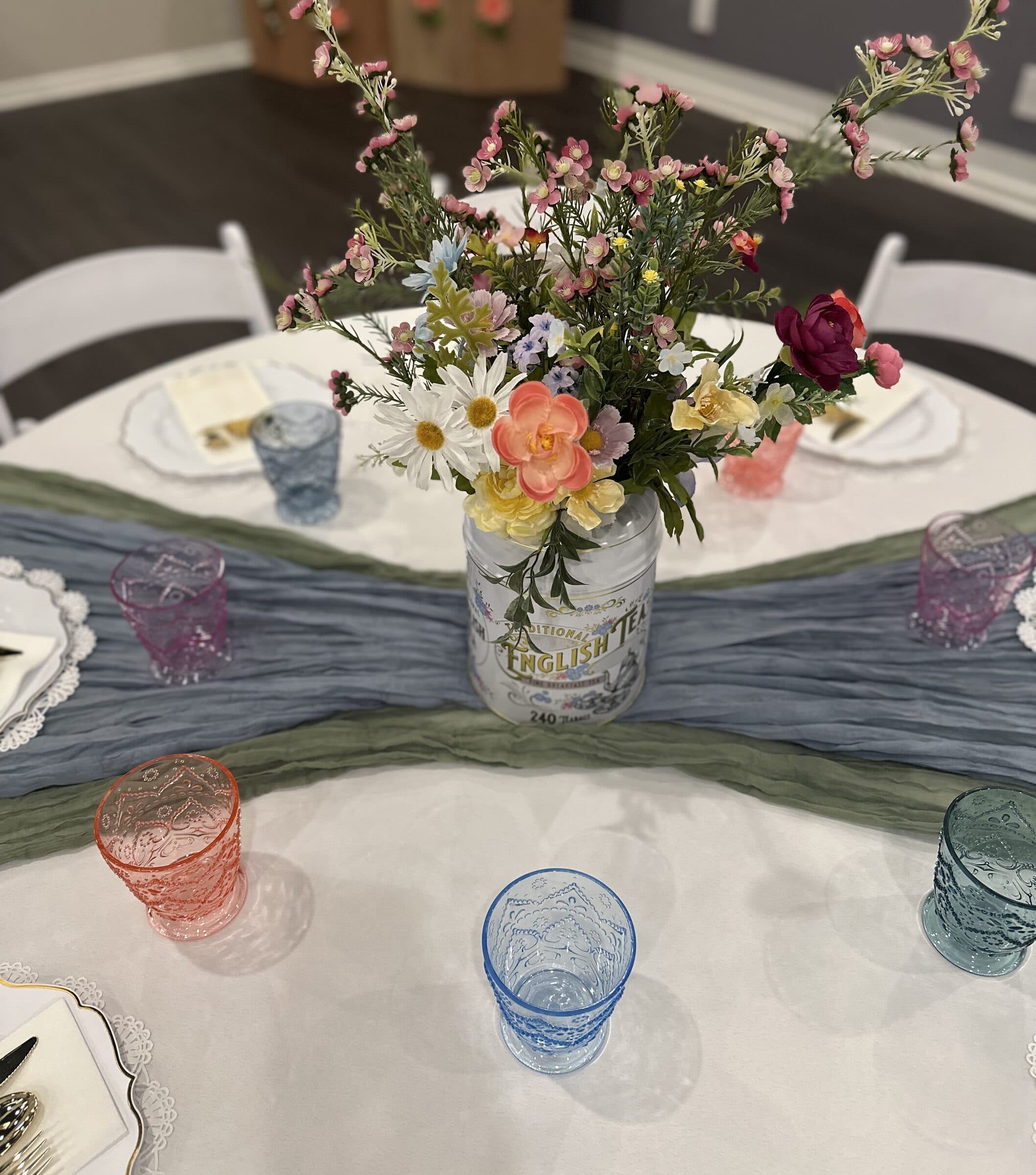 Centerpiece close-up: an English tea tin of pink, yellow and white blooms above layered blue and green runners and pastel goblets, bridal tea at The Cove at St. Johns, Fruit Cove FL