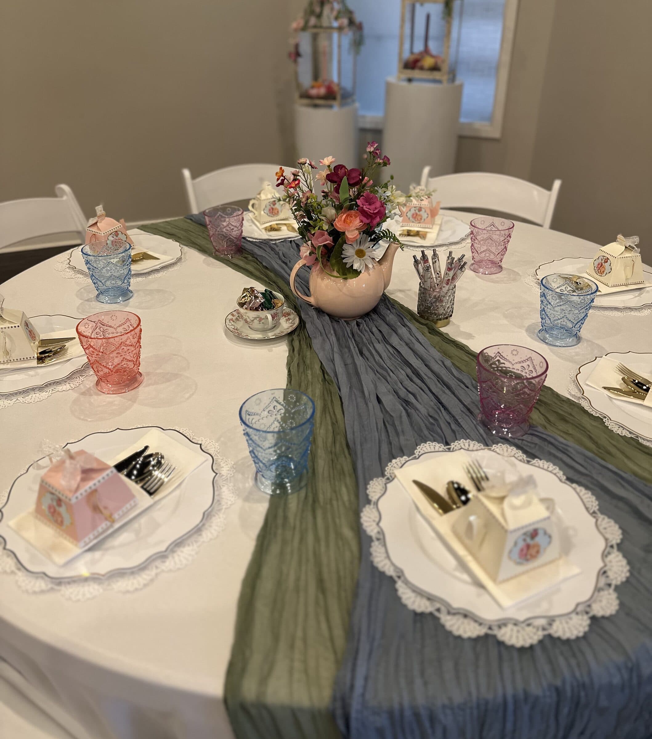 Place settings with teapot-shaped favor boxes on lace-trimmed chargers, gold flatware and a pink teapot of wildflowers at a bridal tea at The Cove at St. Johns, St. Johns County FL