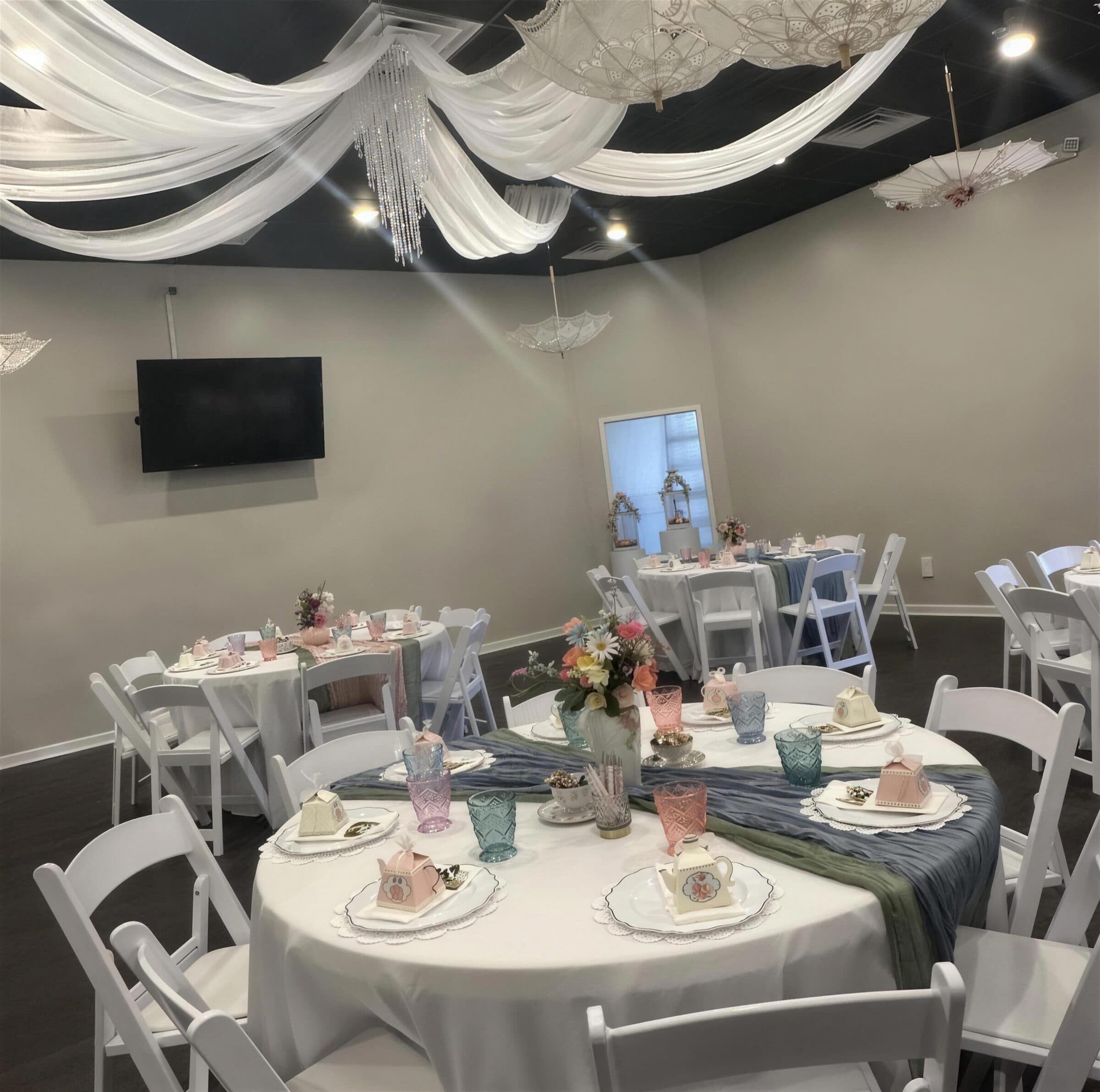 Full room of round tea tables under white ceiling draping, a crystal chandelier and lace parasols hung overhead at a bridal tea at The Cove at St. Johns, Fruit Cove FL