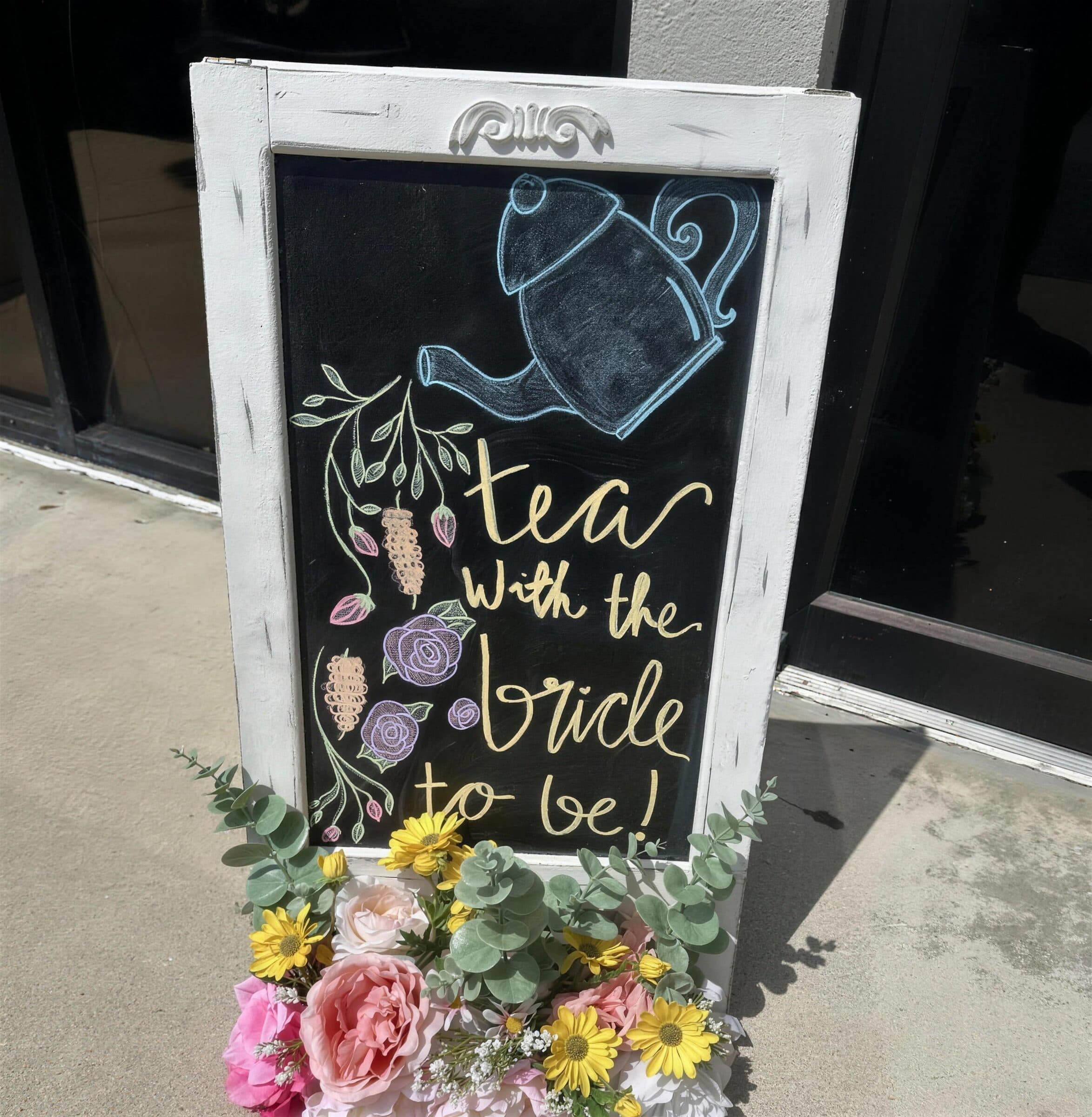 Hand-lettered chalkboard sign reading Tea with the bride to be, in a white frame trimmed with pink and yellow silk flowers, at The Cove at St. Johns, Fruit Cove FL