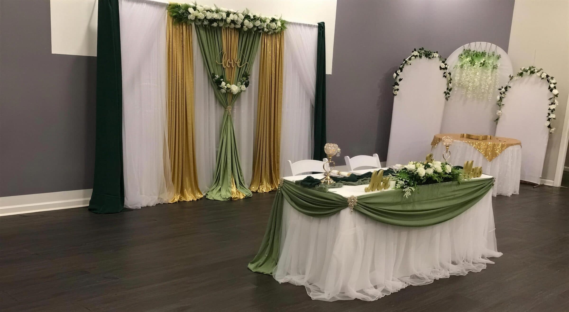 Sweetheart table with sage swag over a white tulle skirt and gold Mr and Mrs letters, backed by emerald, gold and sage drapes and white arch panels at The Cove at St. Johns, near Jacksonville, FL