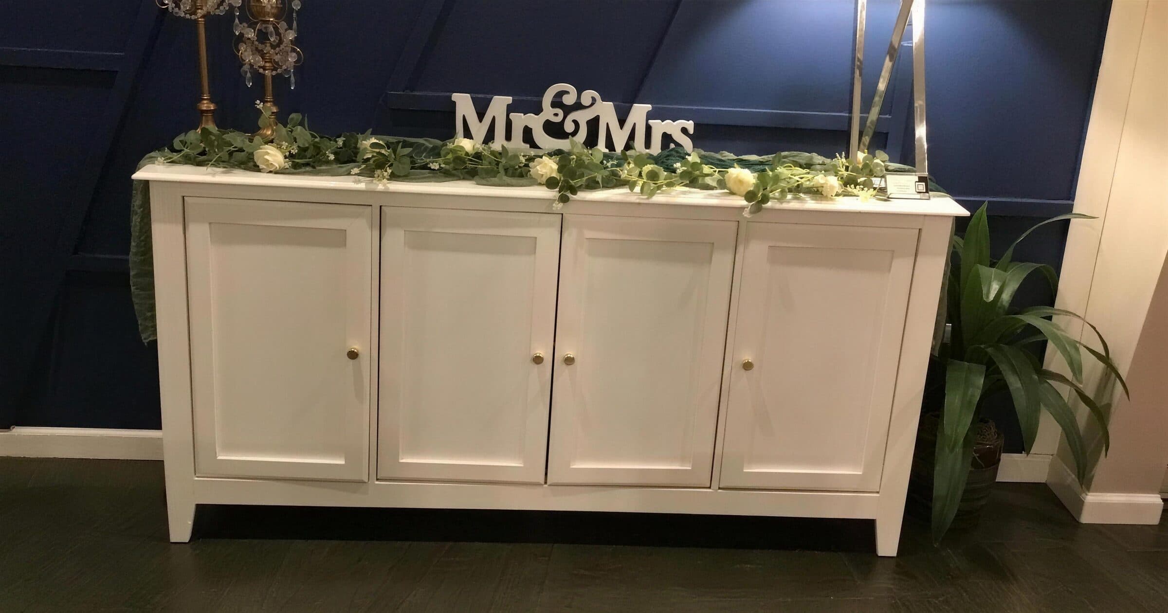 White sideboard styled with a eucalyptus and cream rose garland, a wooden Mr & Mrs sign and crystal candlesticks against the navy panelled wall at The Cove at St. Johns, Fruit Cove, St. Johns County FL