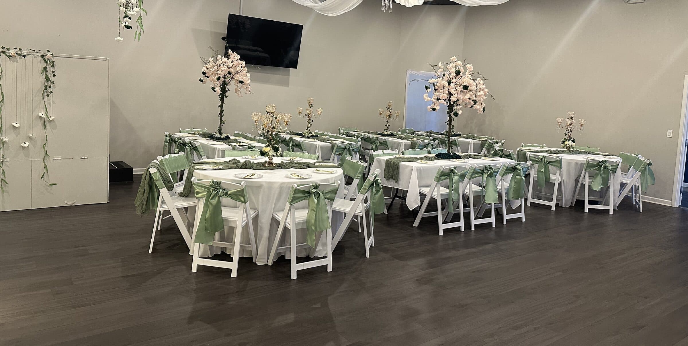 Wide view of the reception room set with round and long tables, sage satin sashes, cheesecloth runners and blush blossom trees under ceiling drapes at The Cove at St. Johns, St. Johns County FL