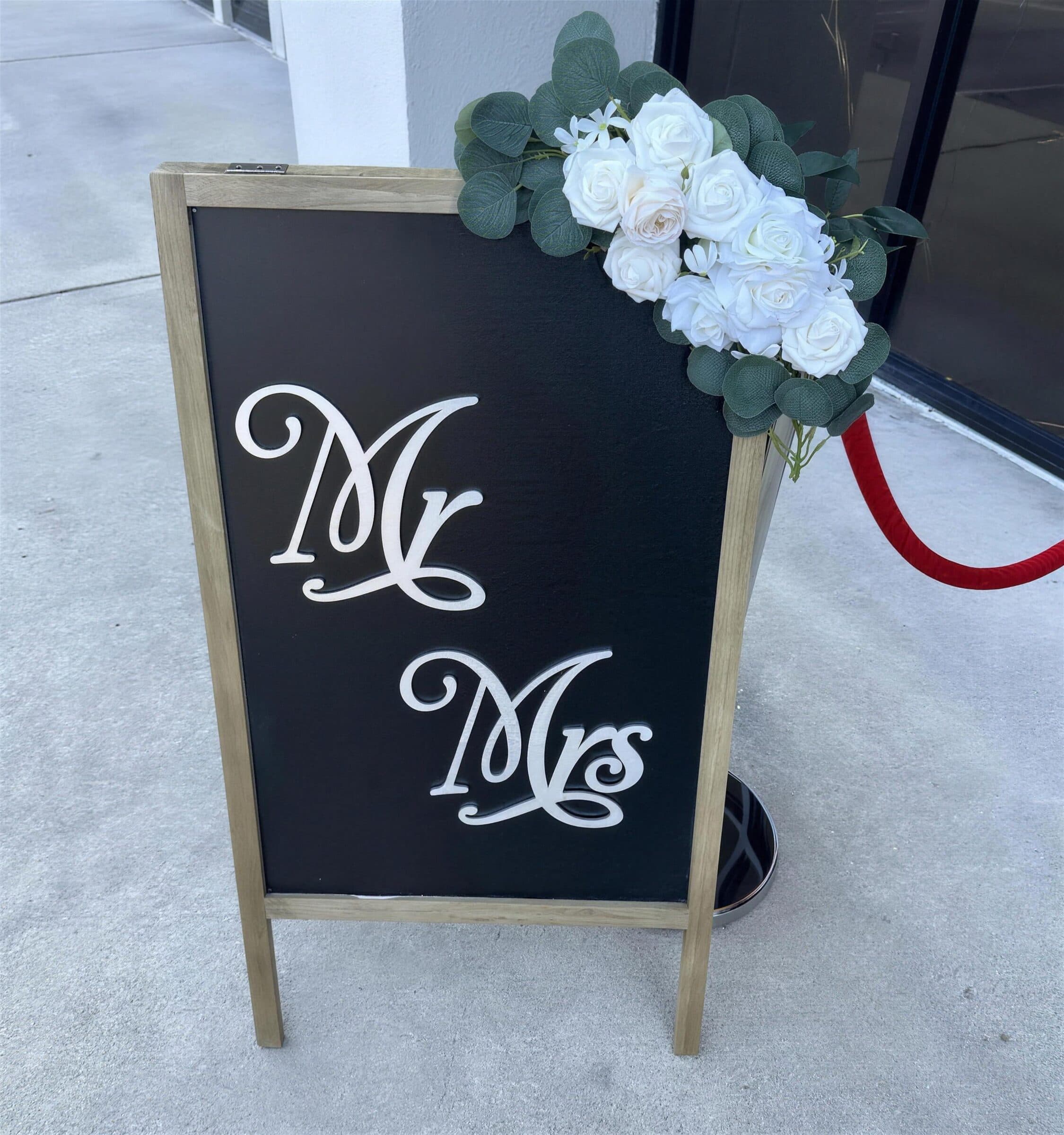 Wooden A-frame chalkboard reading Mr Mrs, topped with a white rose and eucalyptus swag, standing on the walkway outside the entrance at The Cove at St. Johns, Fruit Cove, St. Johns County FL