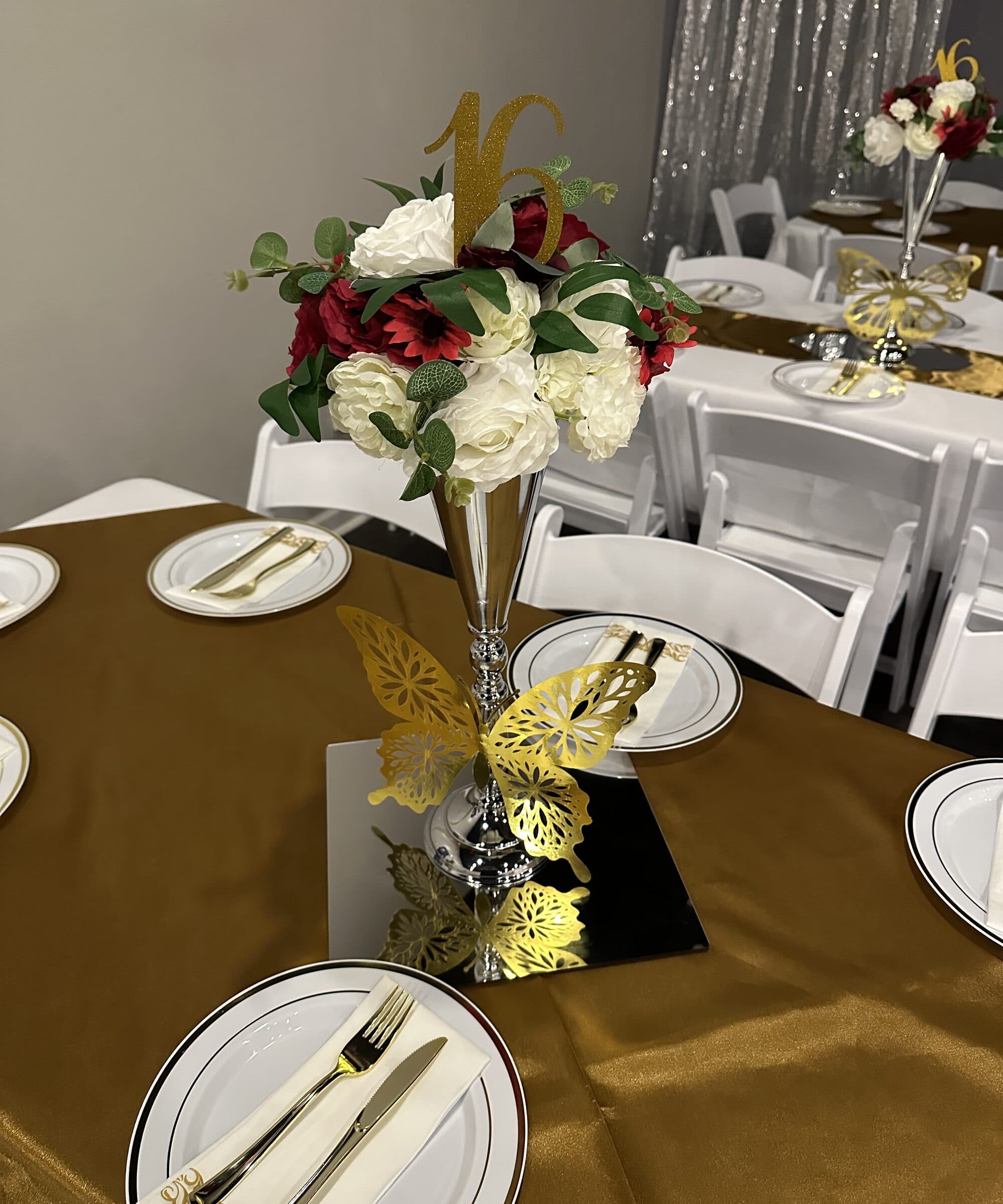 Sweet 16 table in gold satin with a tall silver trumpet vase of red and white roses under a glitter 16 topper, gold paper butterflies and a silver sequin backdrop at The Cove at St. Johns, Fruit Cove FL