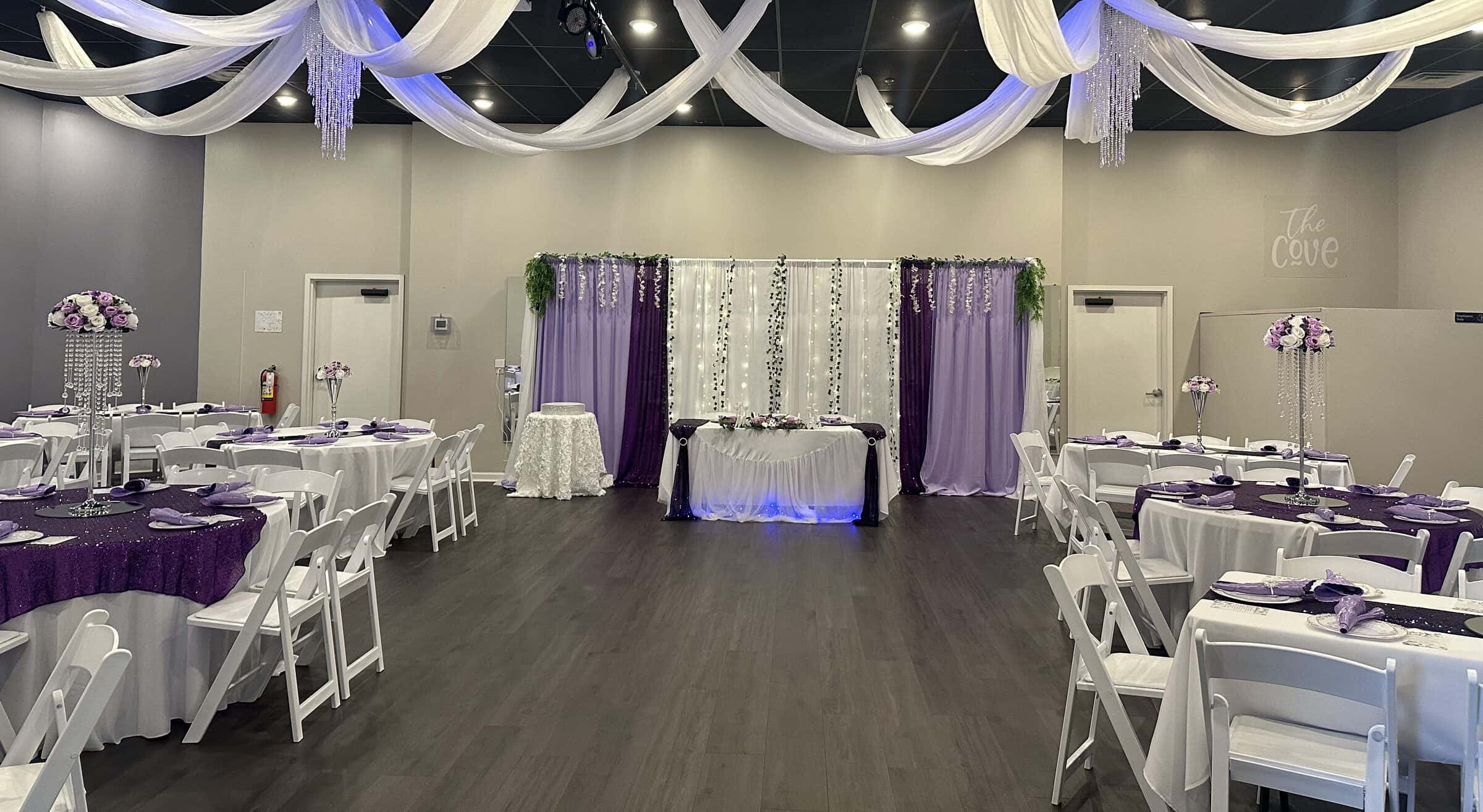 Sweetheart table against a plum, lavender and fairy-light drape wall with trailing greenery, plum sequin guest tables and lilac rose towers at The Cove at St. Johns, near Jacksonville, FL